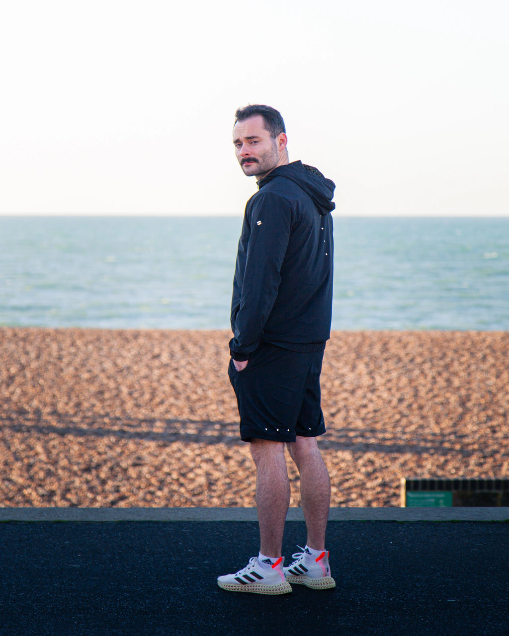 Man standing on a beach wearing a black Satus Core Jacket and Shorts with a blurred sea and sky