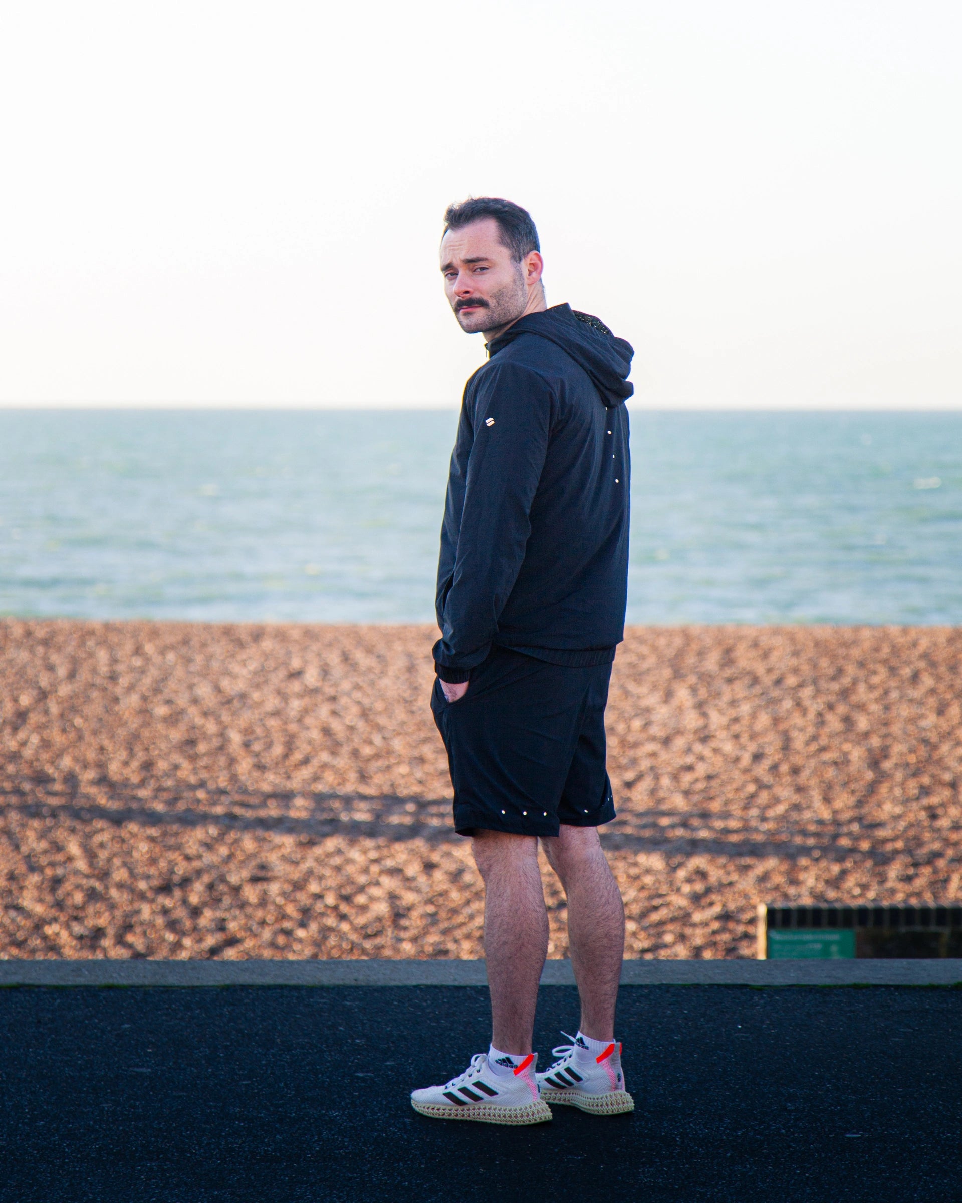 Man standing on a beach wearing a black Satus Core Jacket and Shorts with a blurred sea and sky