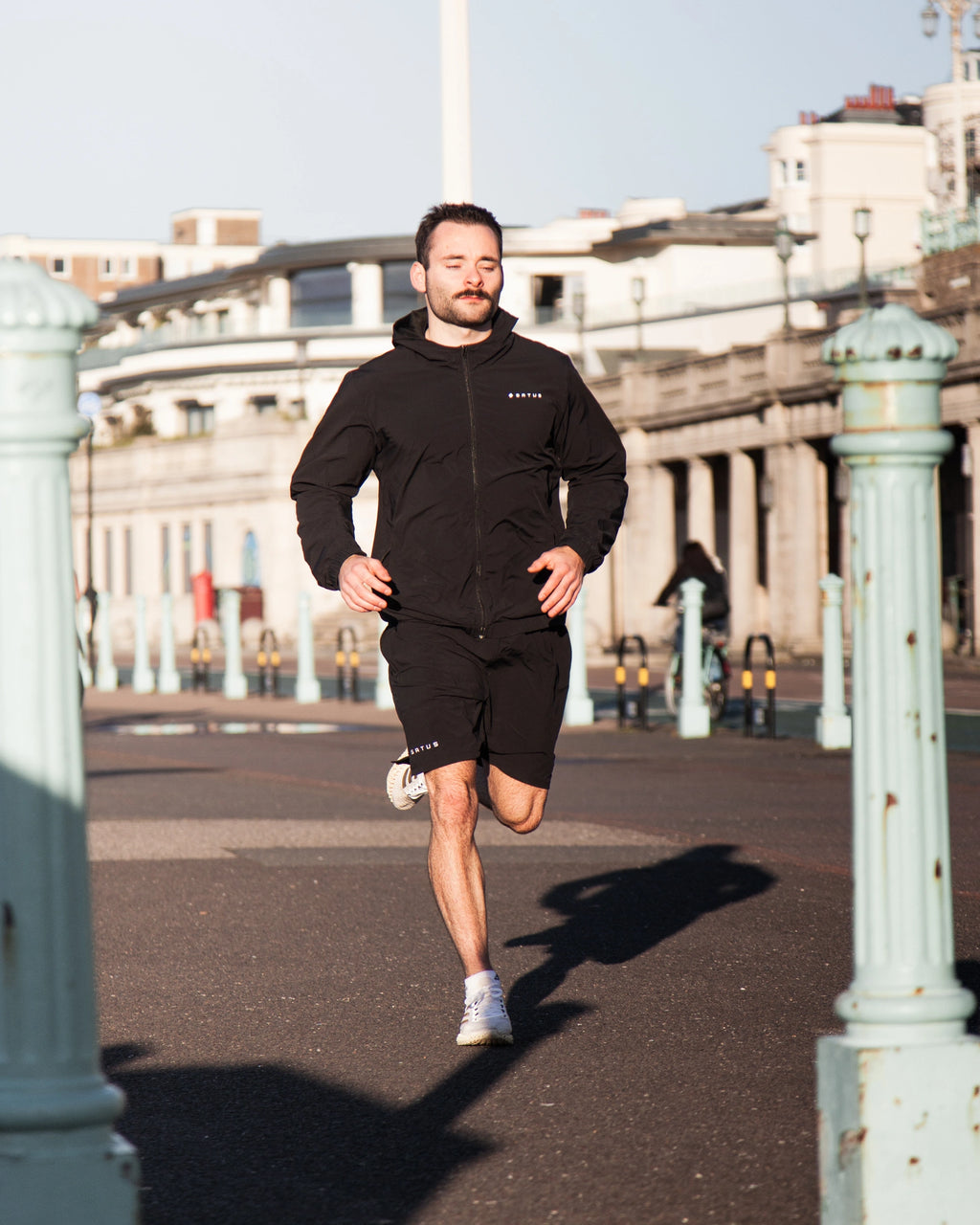 Man running along a sidewalk wearing a black Satus Core Jacket and Shorts