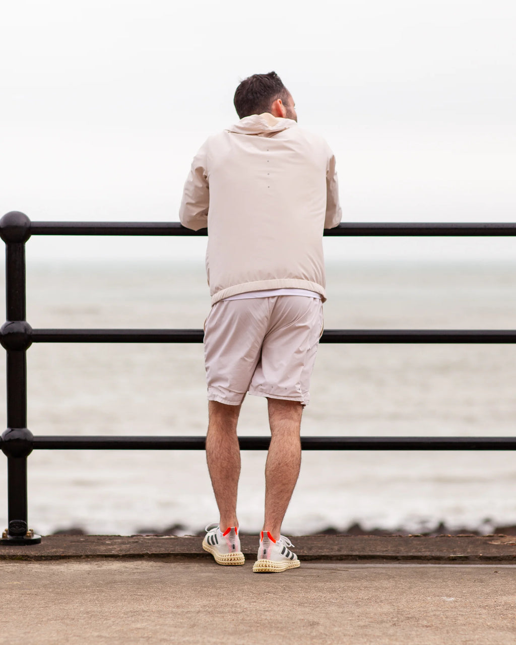 Man standing by a railing looking out at the sea after a run, wearing tan coloured jacket and shots