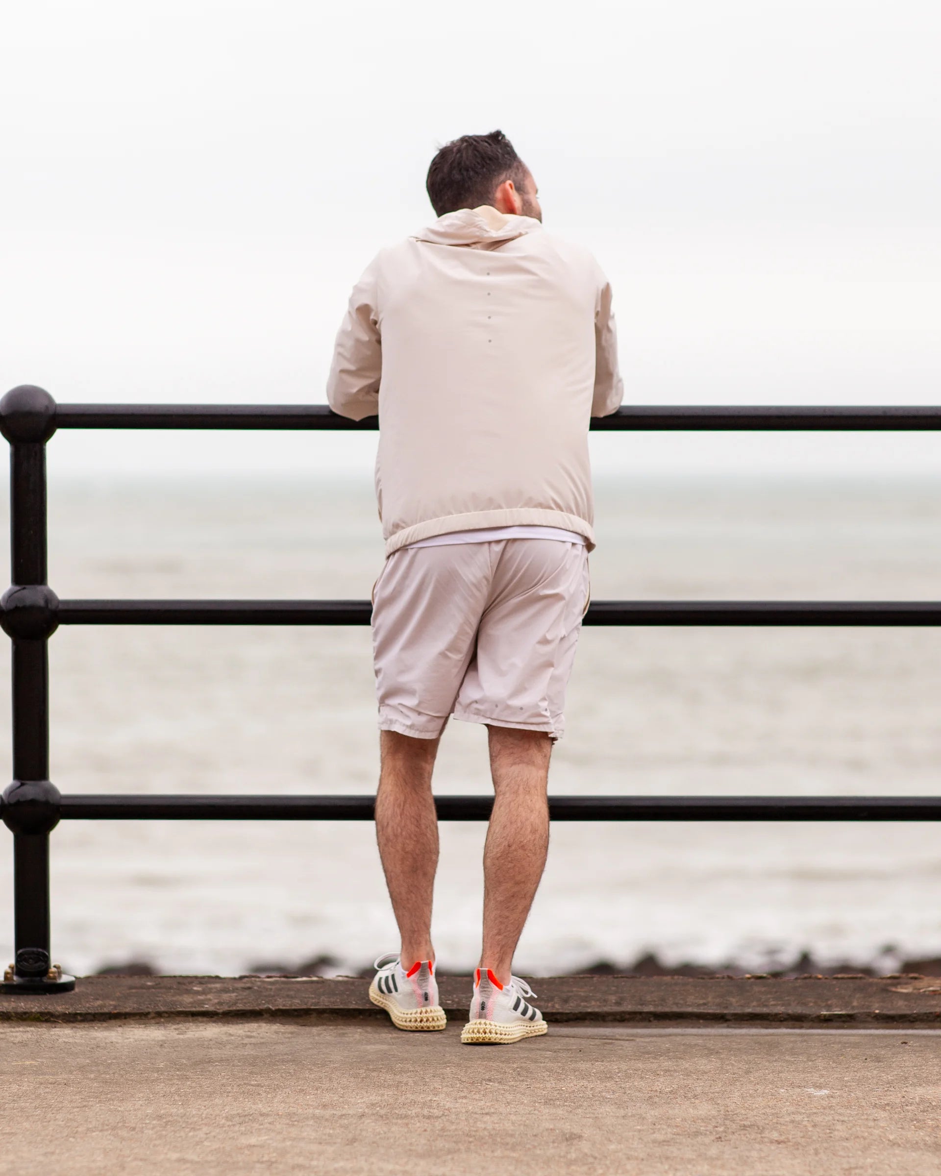 Man standing by a railing looking out at the sea after a run, wearing tan coloured jacket and shots