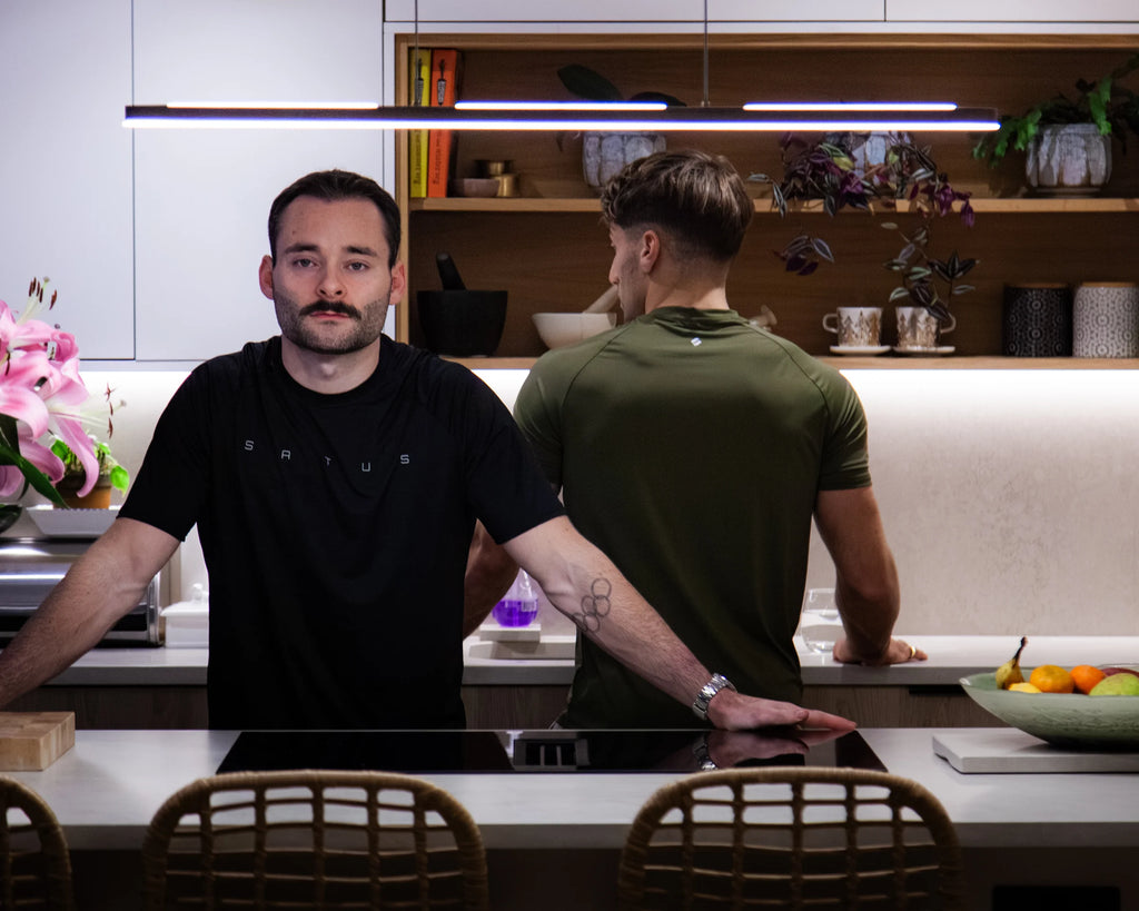 Two men standing in a modern kitchen with shelves and decor, wearing sportswear.