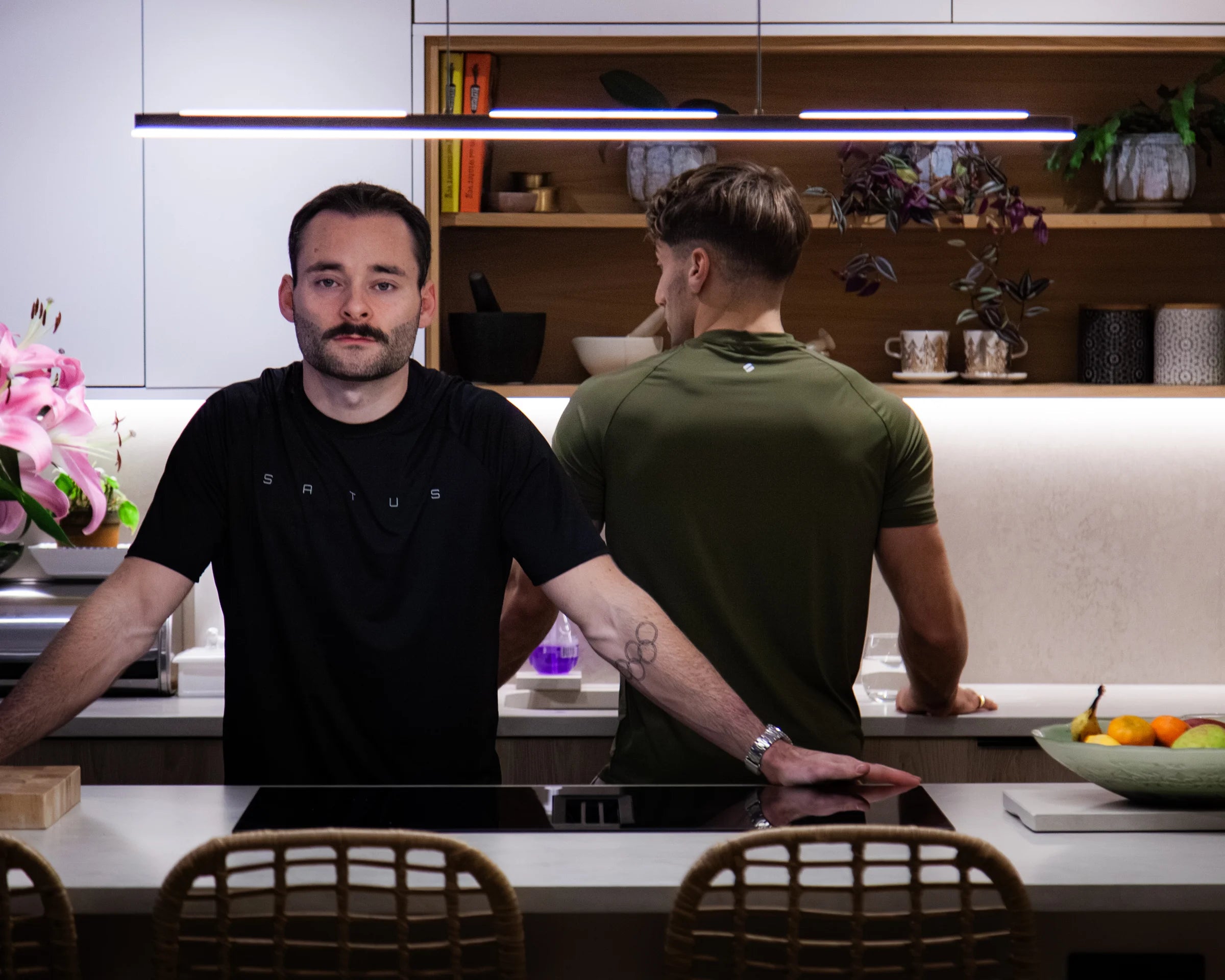 Two men standing in a modern kitchen with shelves and decor, wearing sportswear.