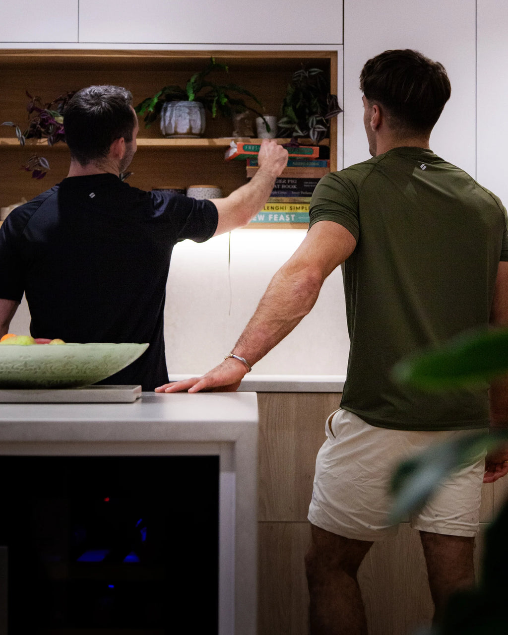 Two men in a kitchen, one reaching into an open cabinet wearing sports clothing
