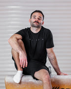 Man sitting on a gymnastics pommel horse wearing a black sports t-shirt and shorts with olympic rings tattoos.