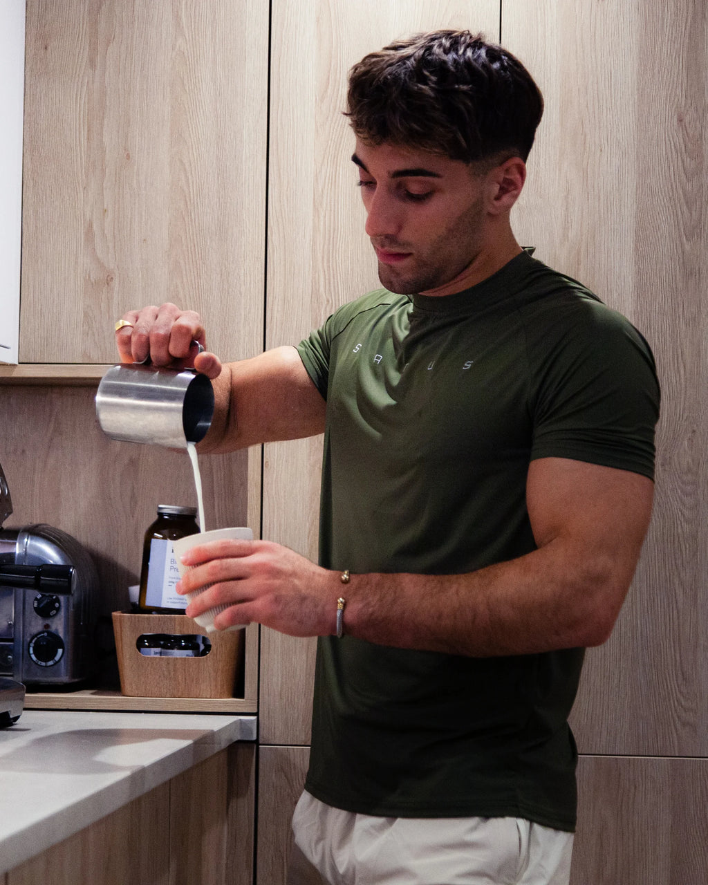 Man in a kitchen pouring a coffee into a cup wearing olive sports top