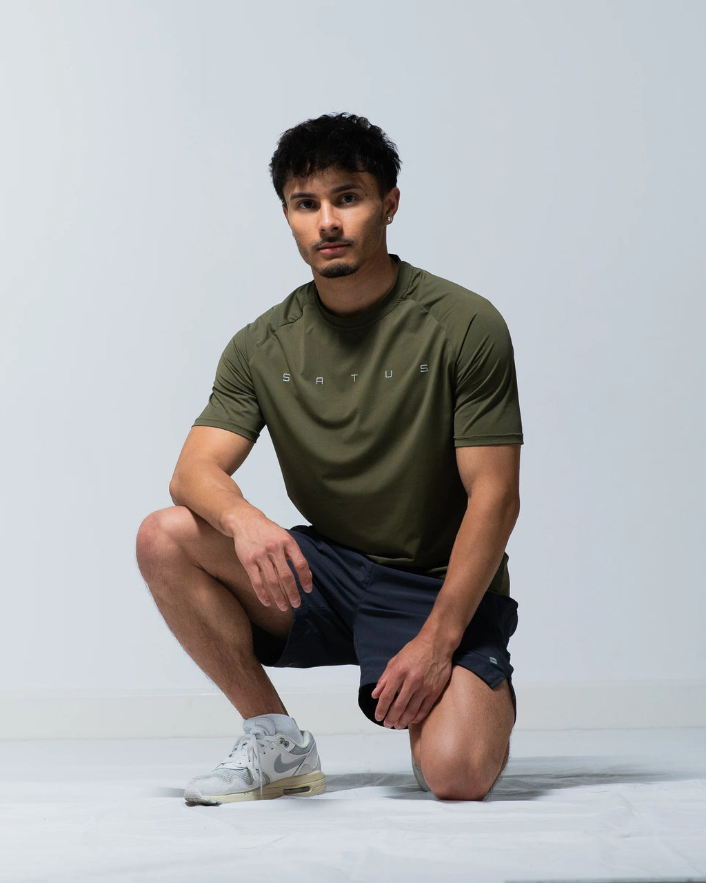 Man wearing an olive green athletic shirt and dark shorts on a light gray background