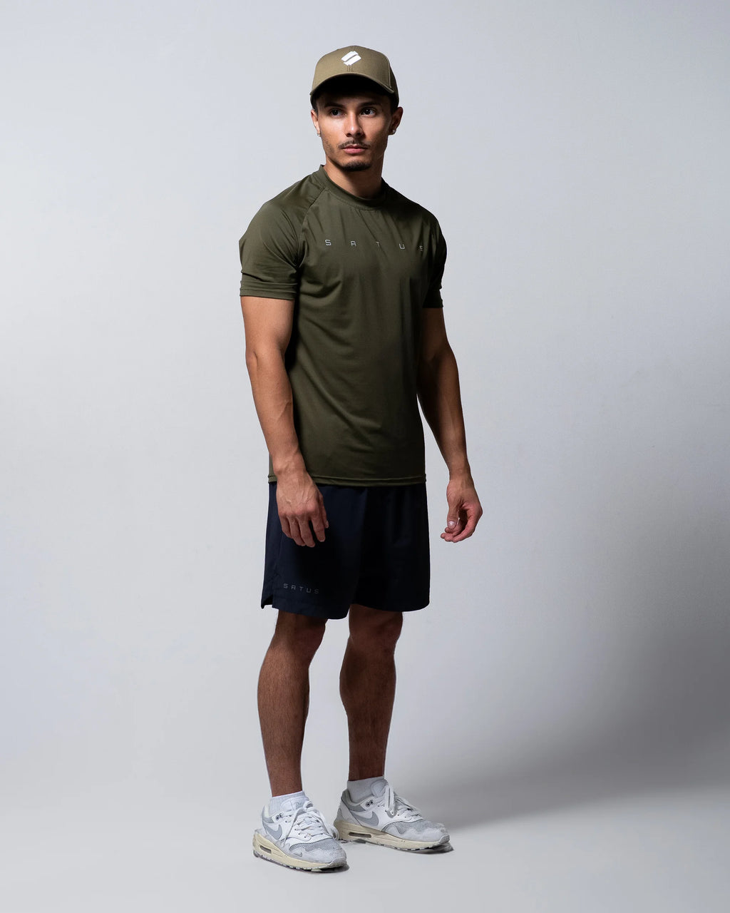 Man wearing an olive green t-shirt, black shorts, and a cap on a white background