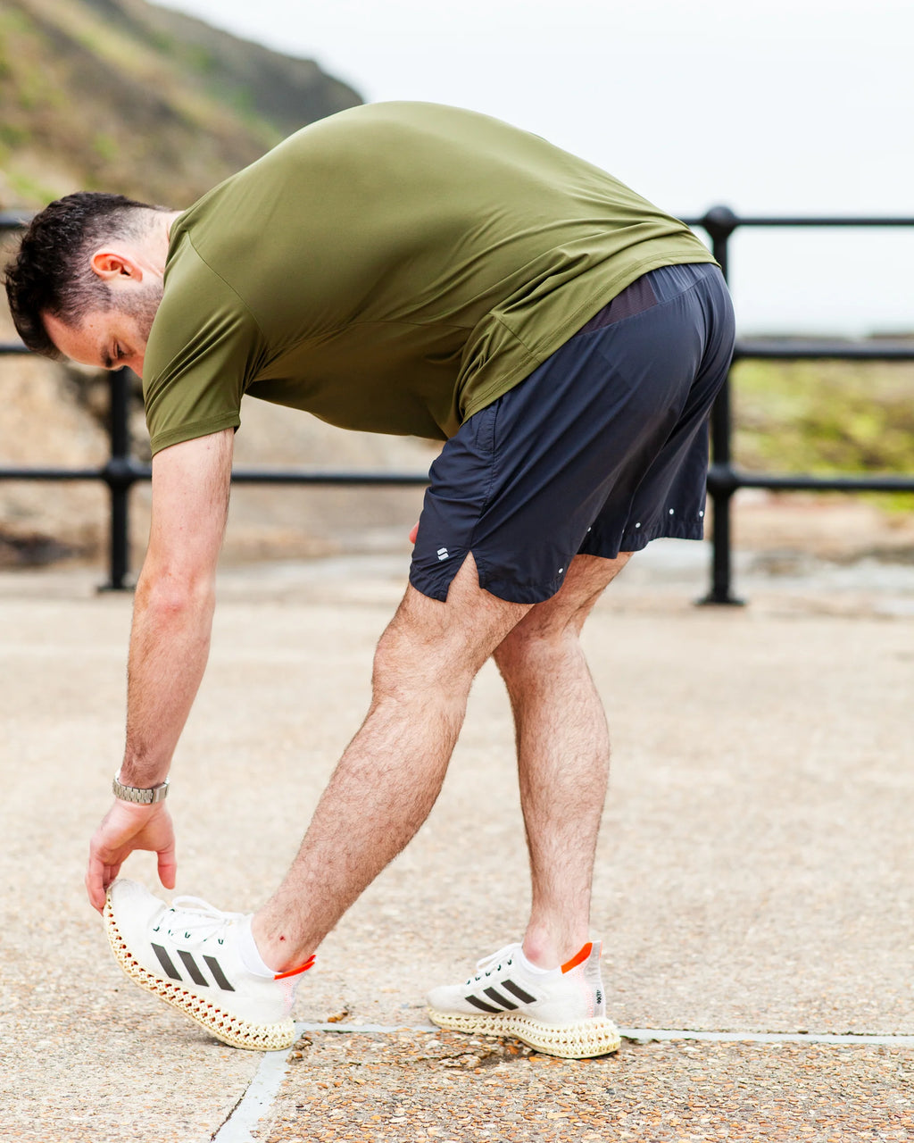 Person stretching outdoors on a path with a scenic background, wearing training shorts and an olive sports t-shirt