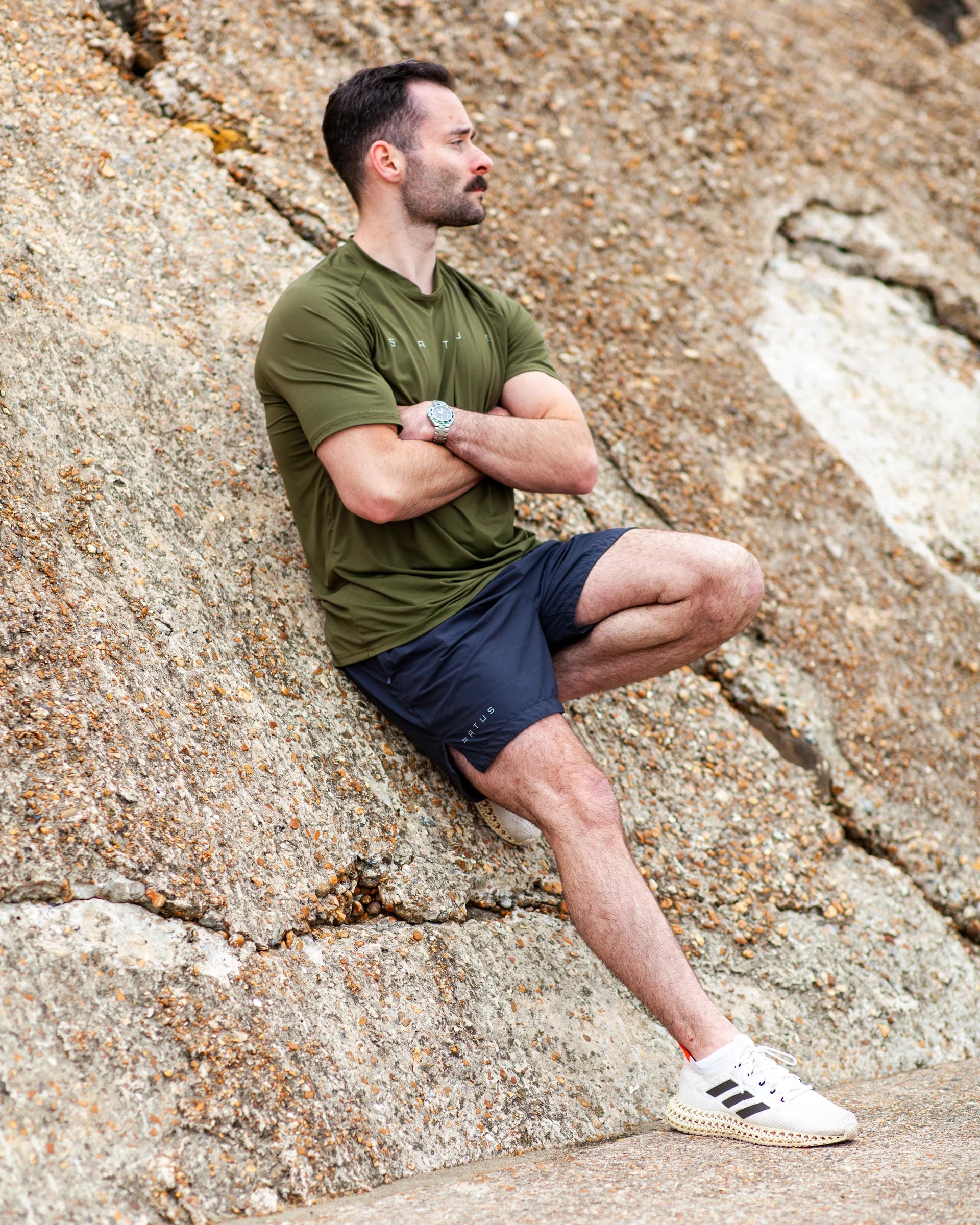 Man sitting on a rocky surface wearing an satus core tee in olive and training shorts.