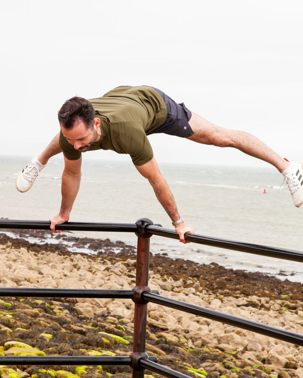Man doing calisthenics on a railing by the beach wearing an olive t-shirt and training shorts