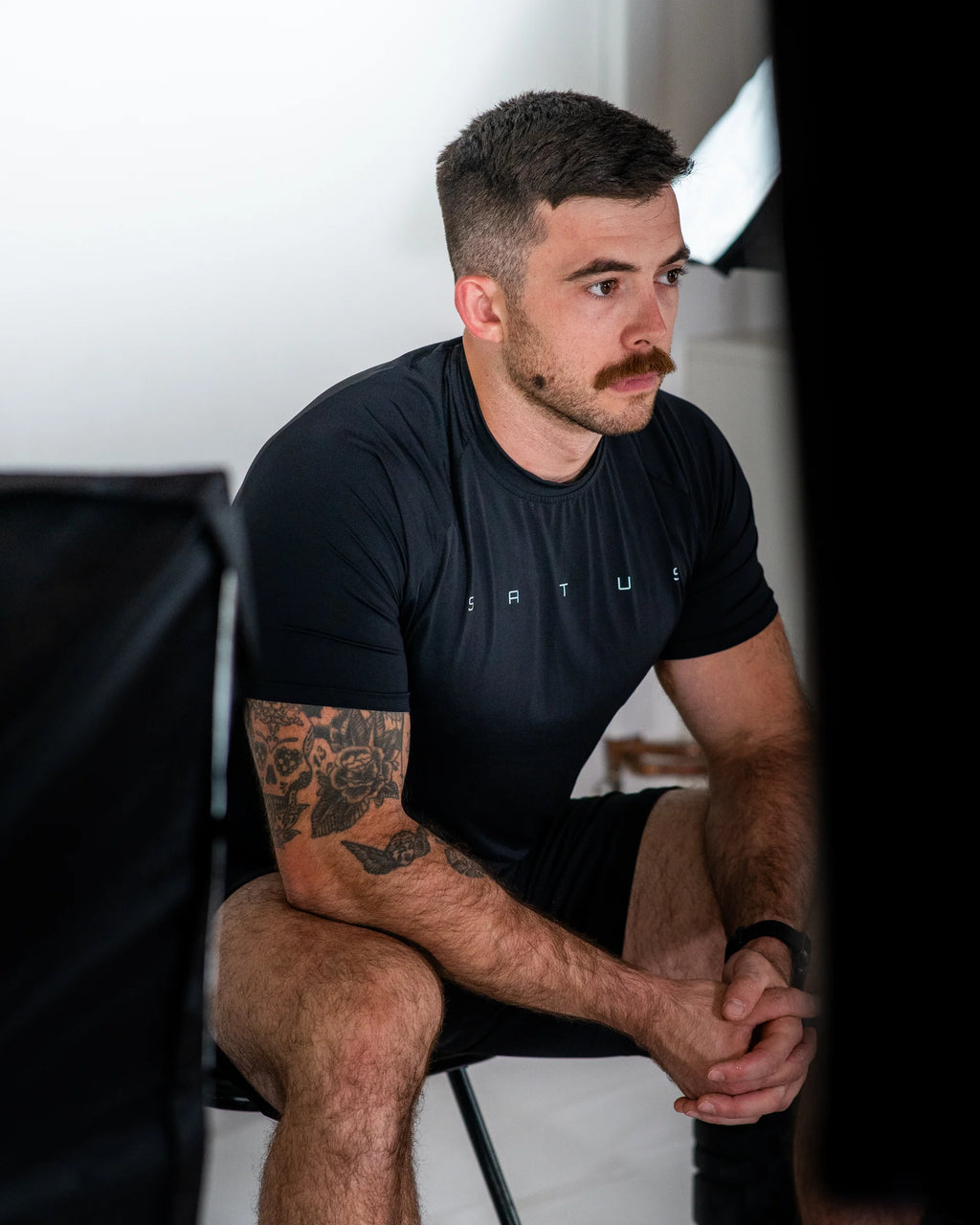 Man wearing a black t-shirt with 'Satus' branding, sitting indoors.