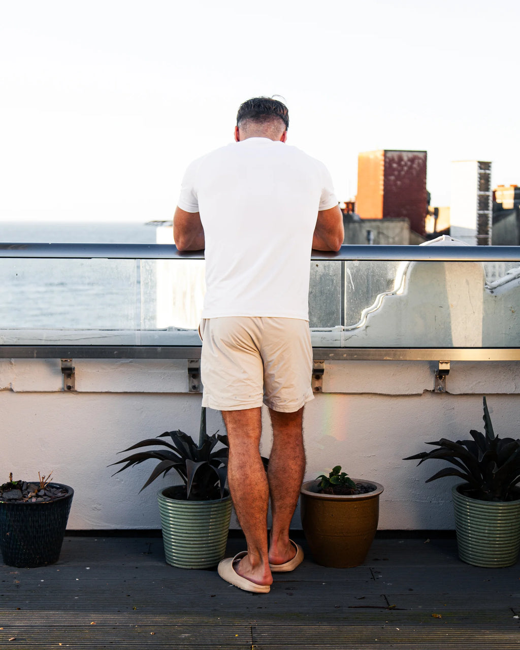 Man standing on a balcony with plants, looking out over a cityscape, wearing white t-shirt and tan shorts