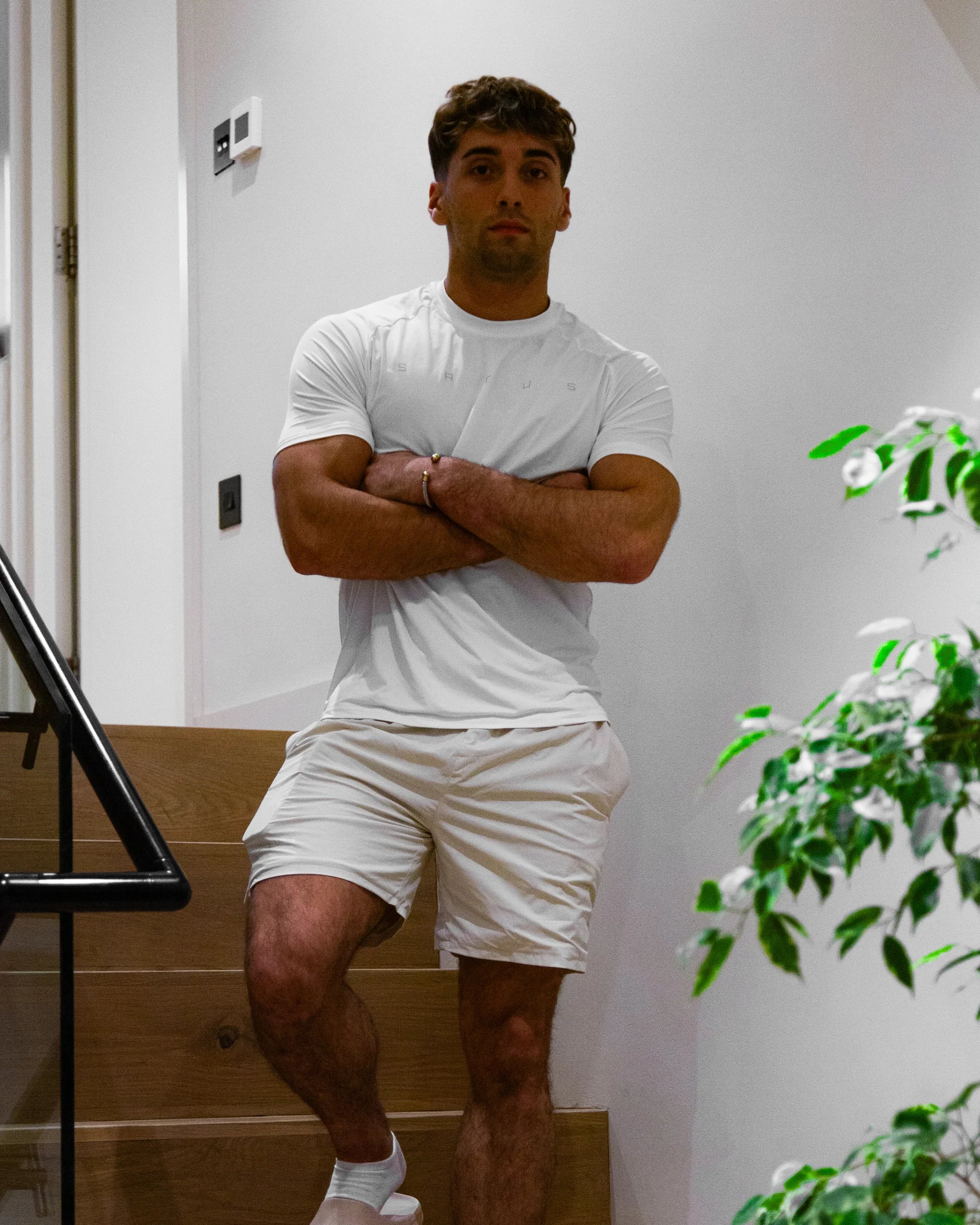 Man in white sports tee t-shirt and shorts standing indoors with a plant in the background