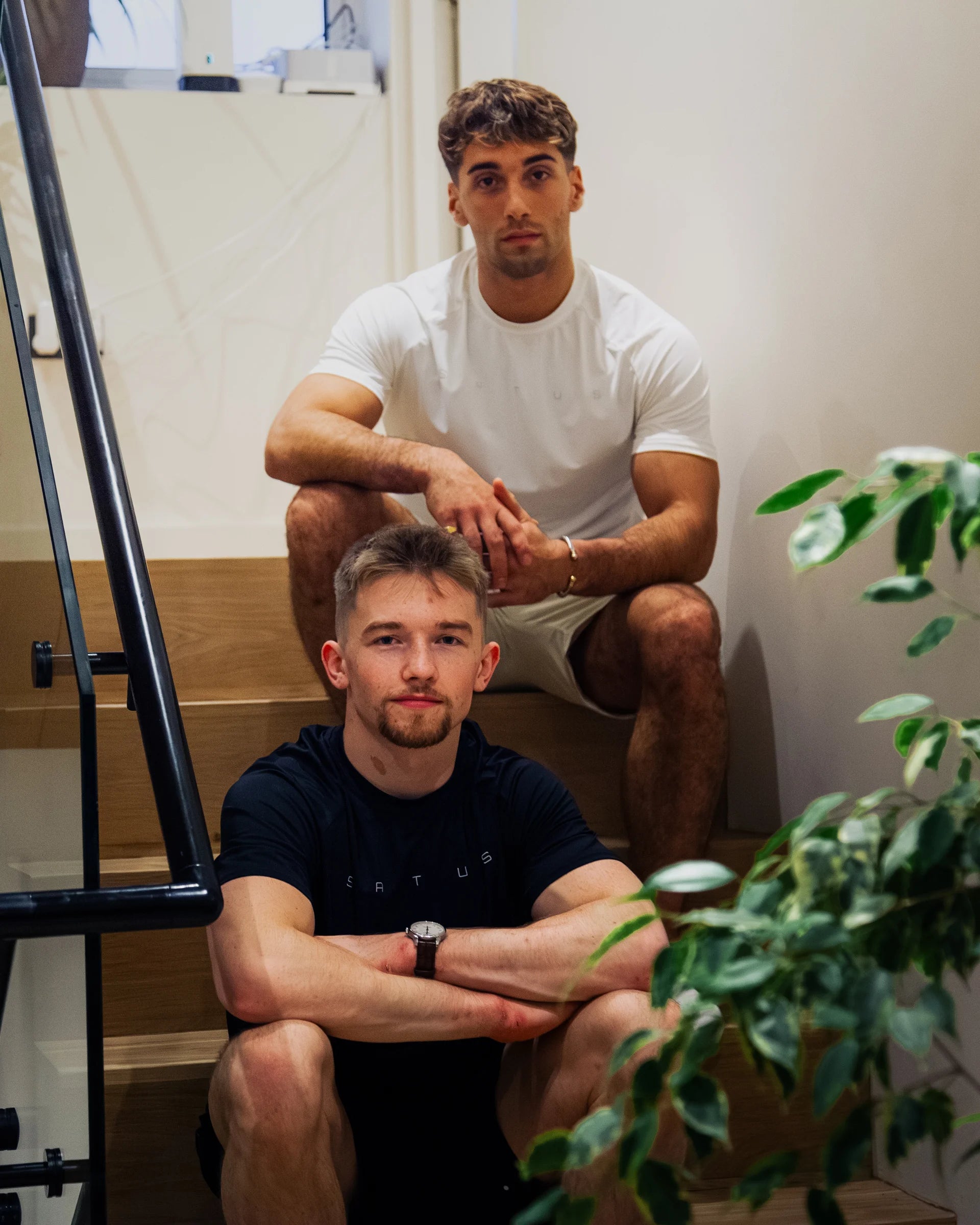Two men sitting on a staircase with a plant in the foreground, wearing sports t-shirts