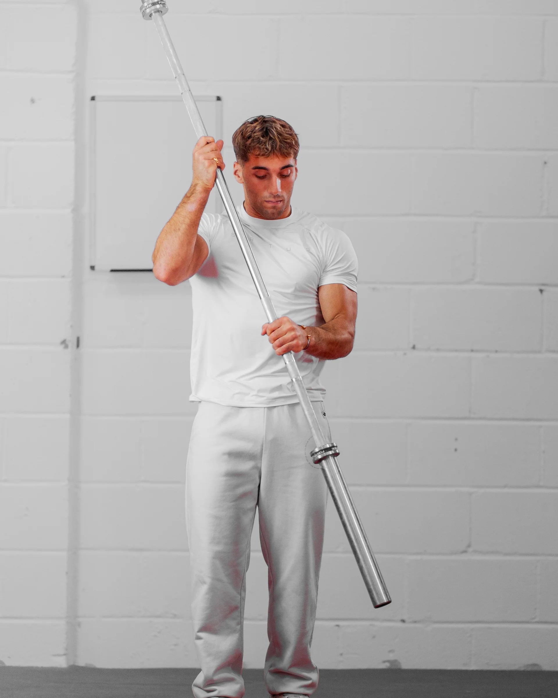 Man holding a barbell against a white brick wall wearing sportswear.
