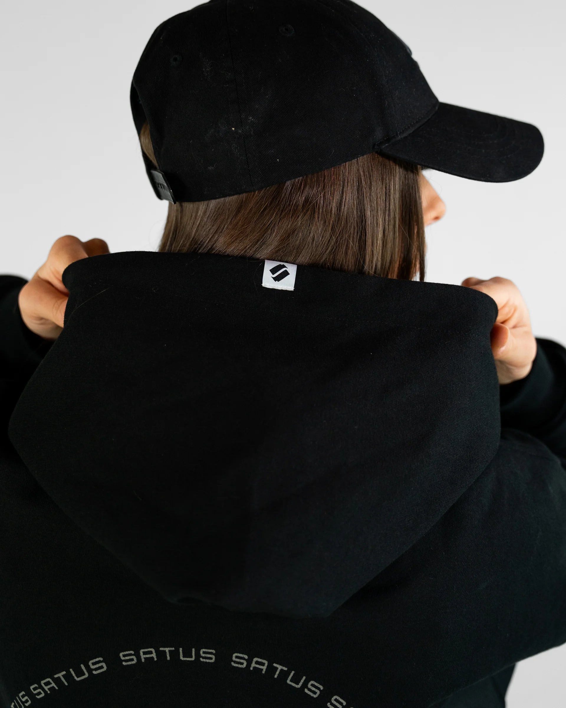 Person wearing a black hoodie and cap with a visible brand logo on a light gray background