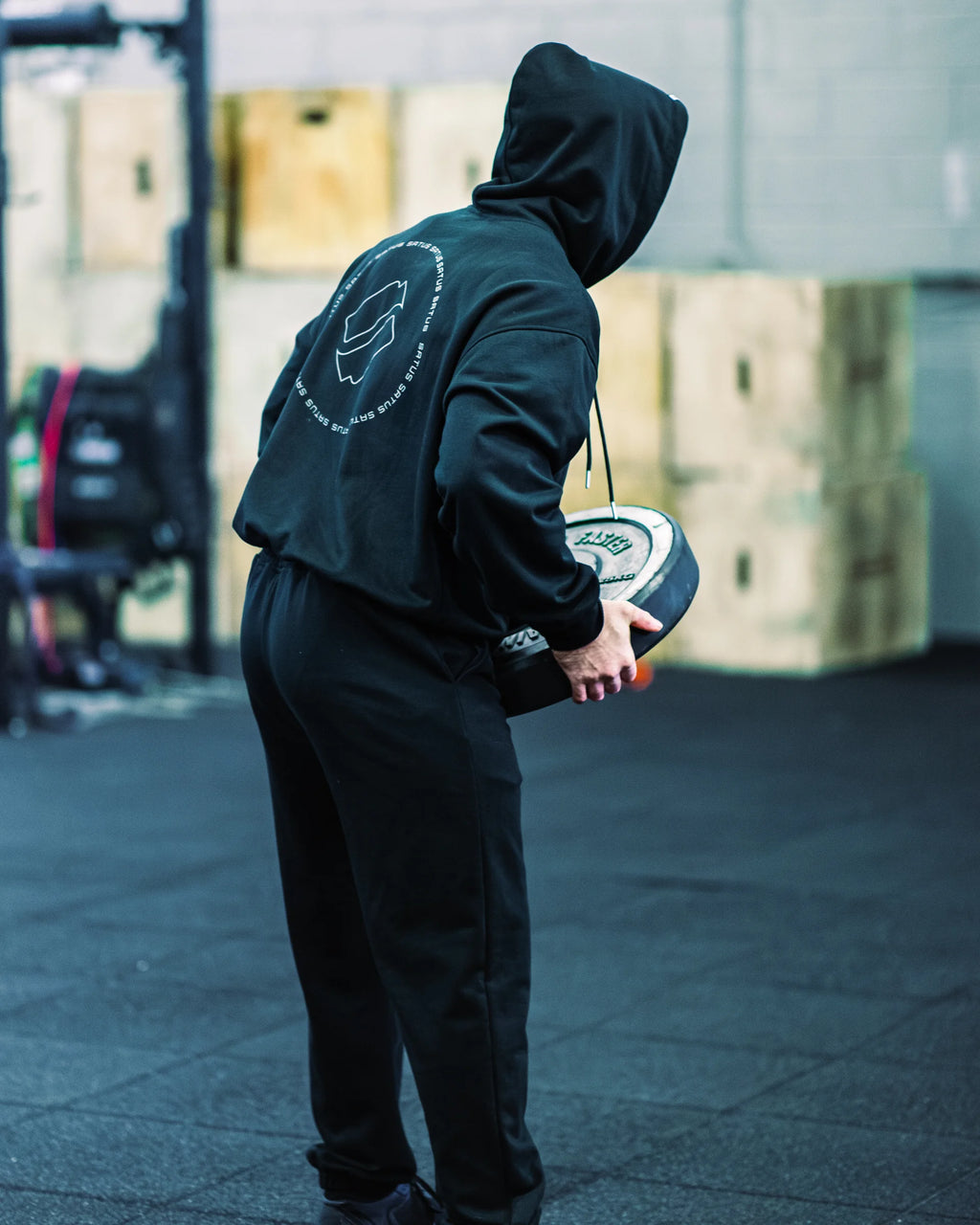 Person wearing a black hoodie with a logo, holding a weight in a gym