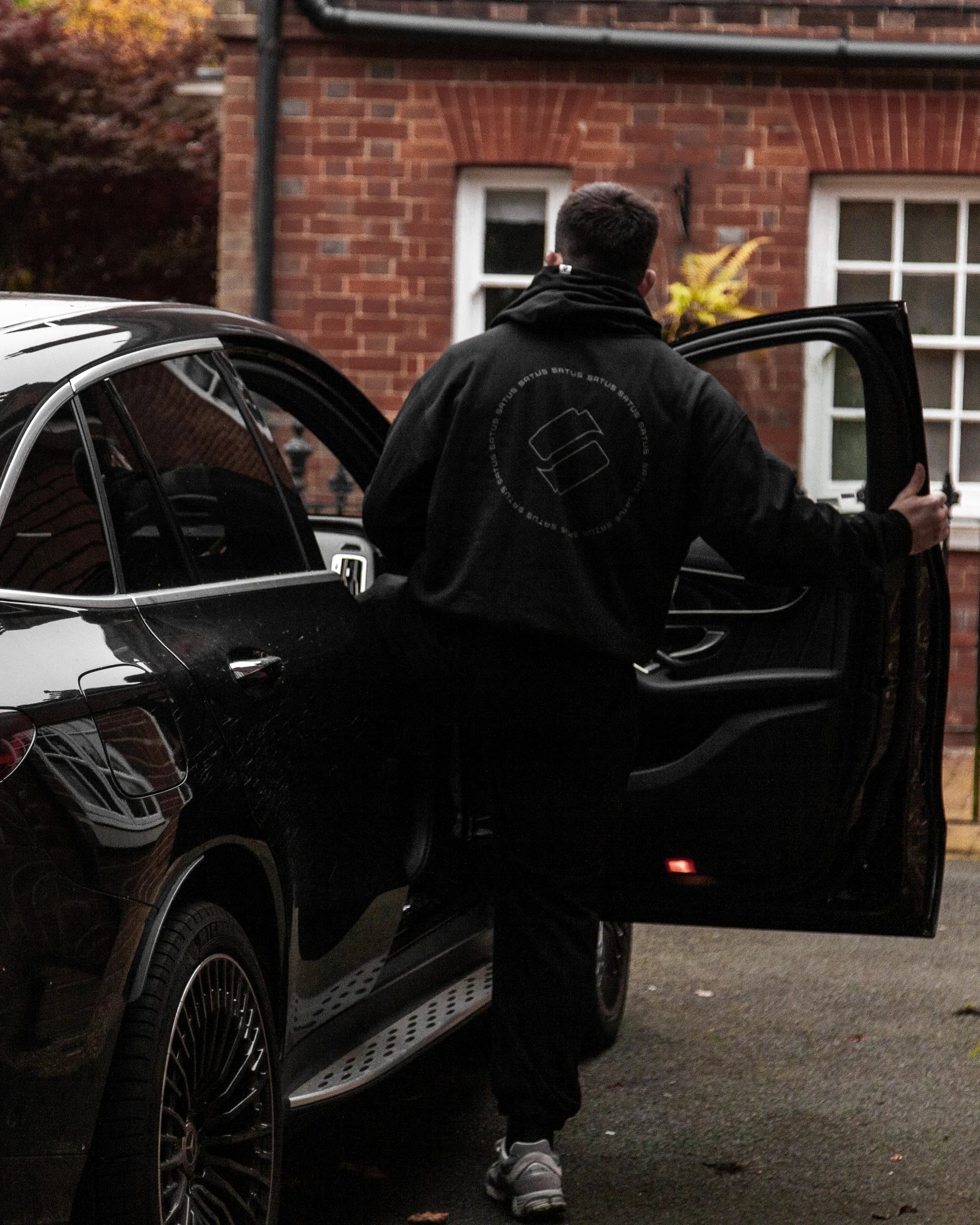 Person opening a car door in front of a brick building wearing premium hoodie and joggers in black