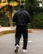 Person wearing a black hoodie and joggers with a minimal logo, walking outdoors.