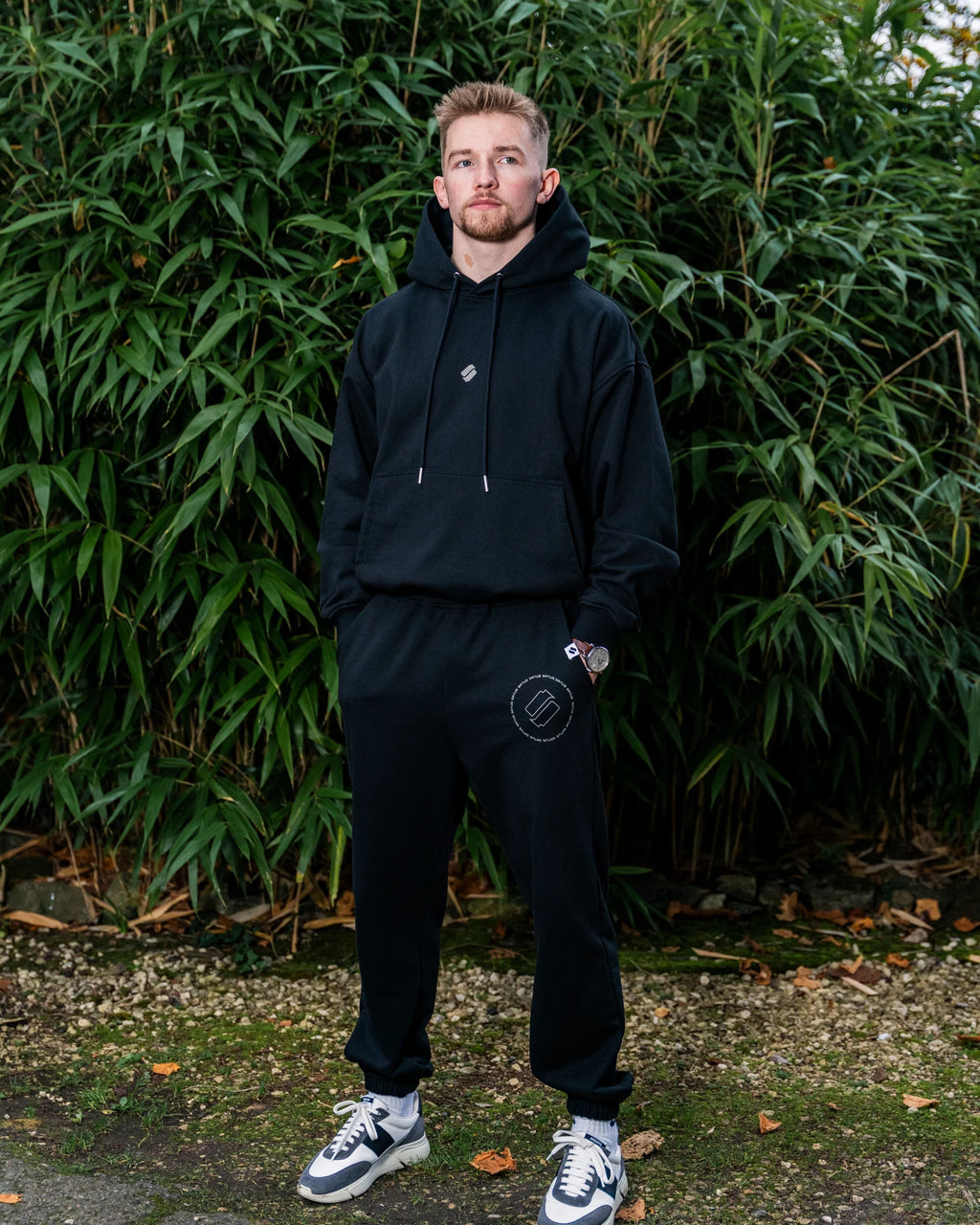 Man wearing a black hoodie and joggers standing in front of green foliage