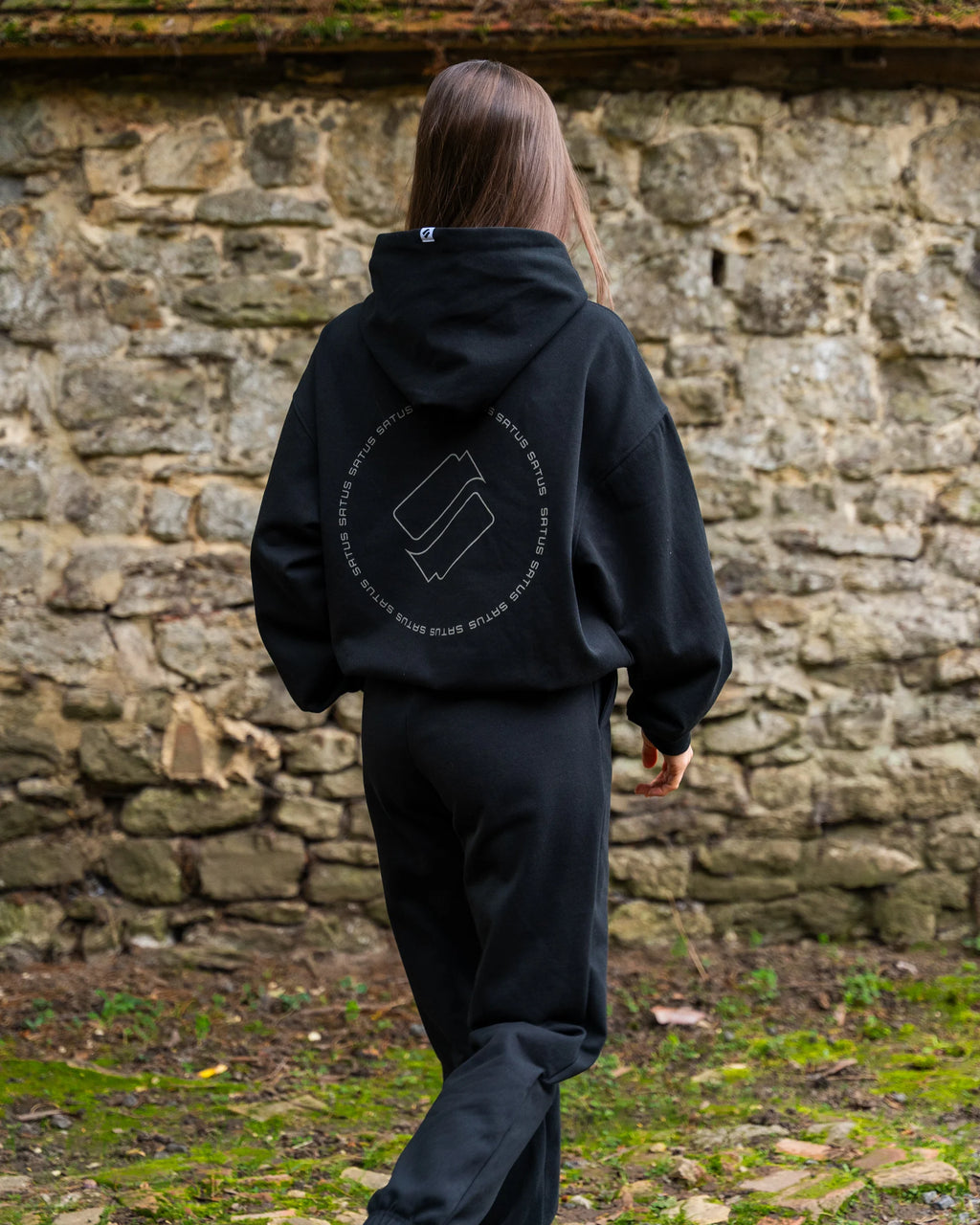 Person wearing a black hoodie with a logo and text, walking in front of a stone wall.