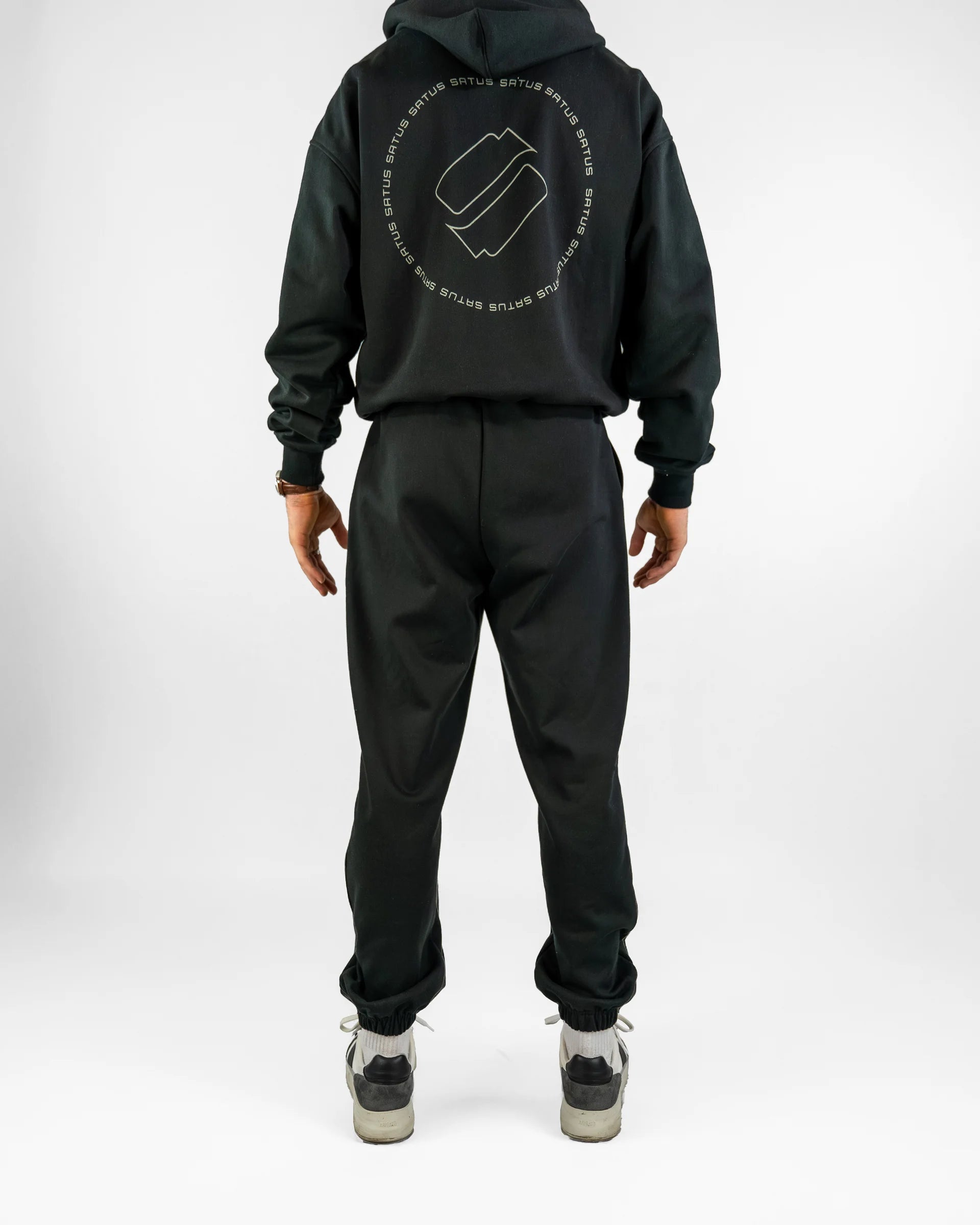 Person wearing a black hoodie and joggers set with a logo on the back, standing against a white background.