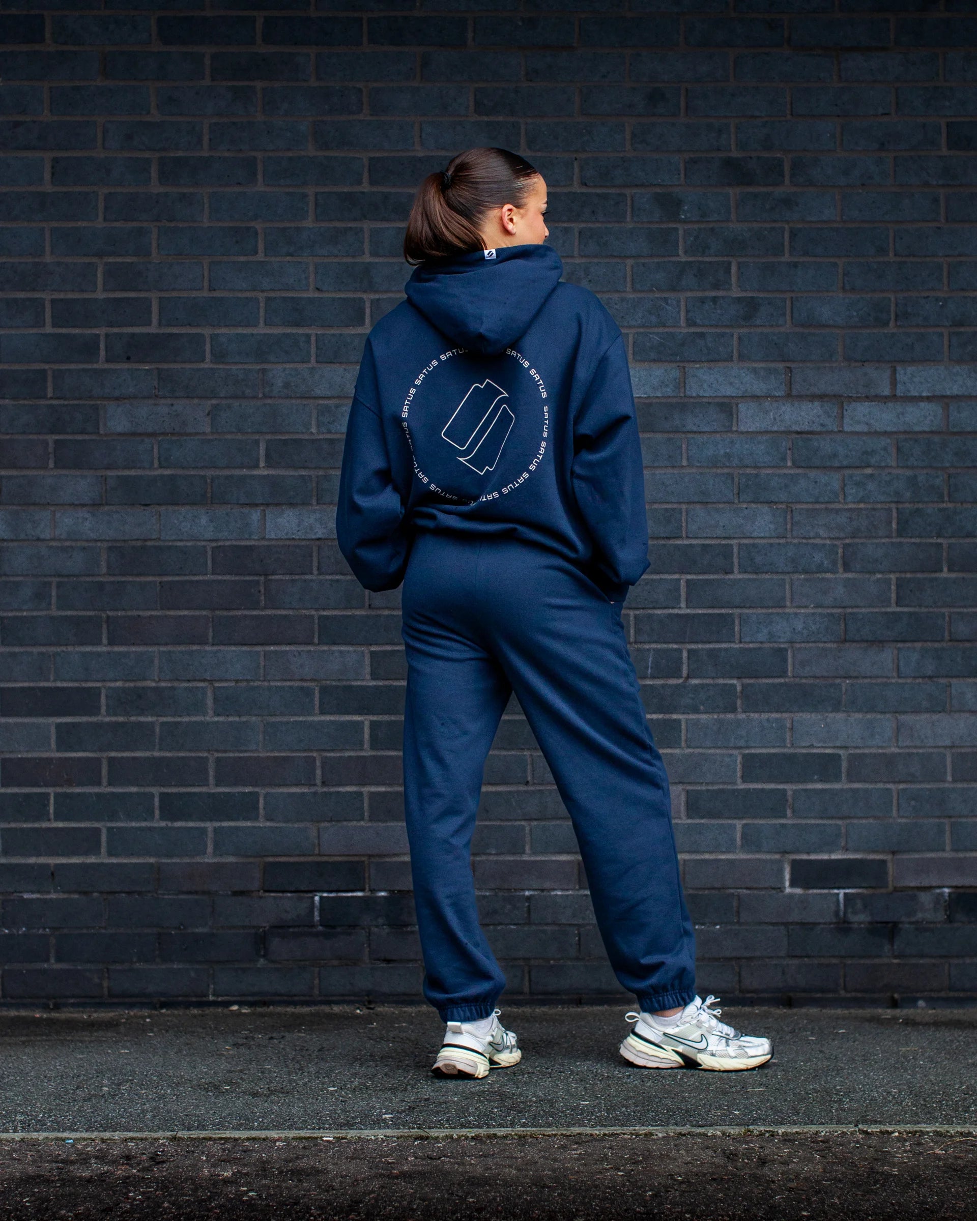 Girl wearing a blue tracksuit with a logo on the back, standing against a dark brick wall.