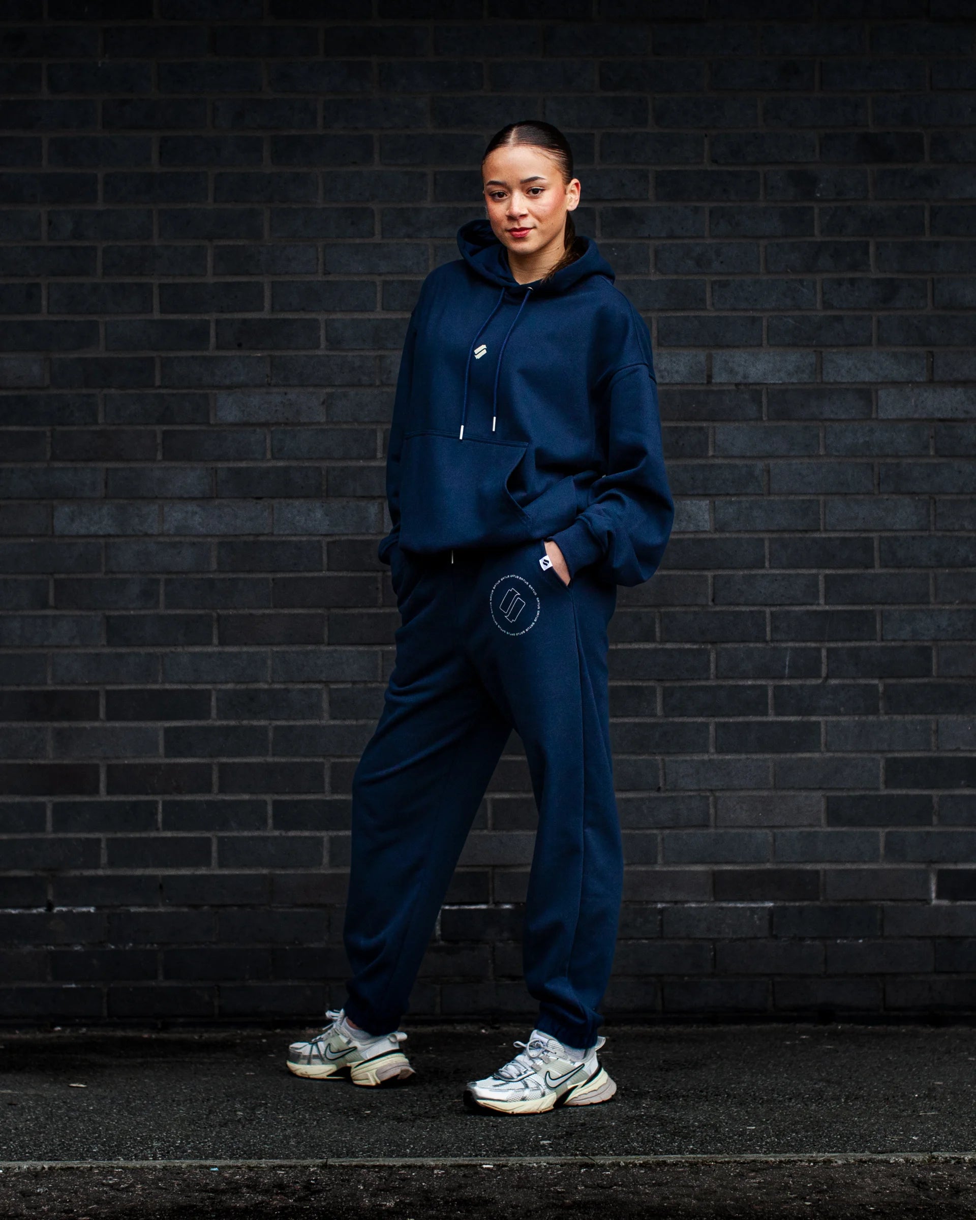 Person wearing a navy tracksuit standing against a dark brick wall.