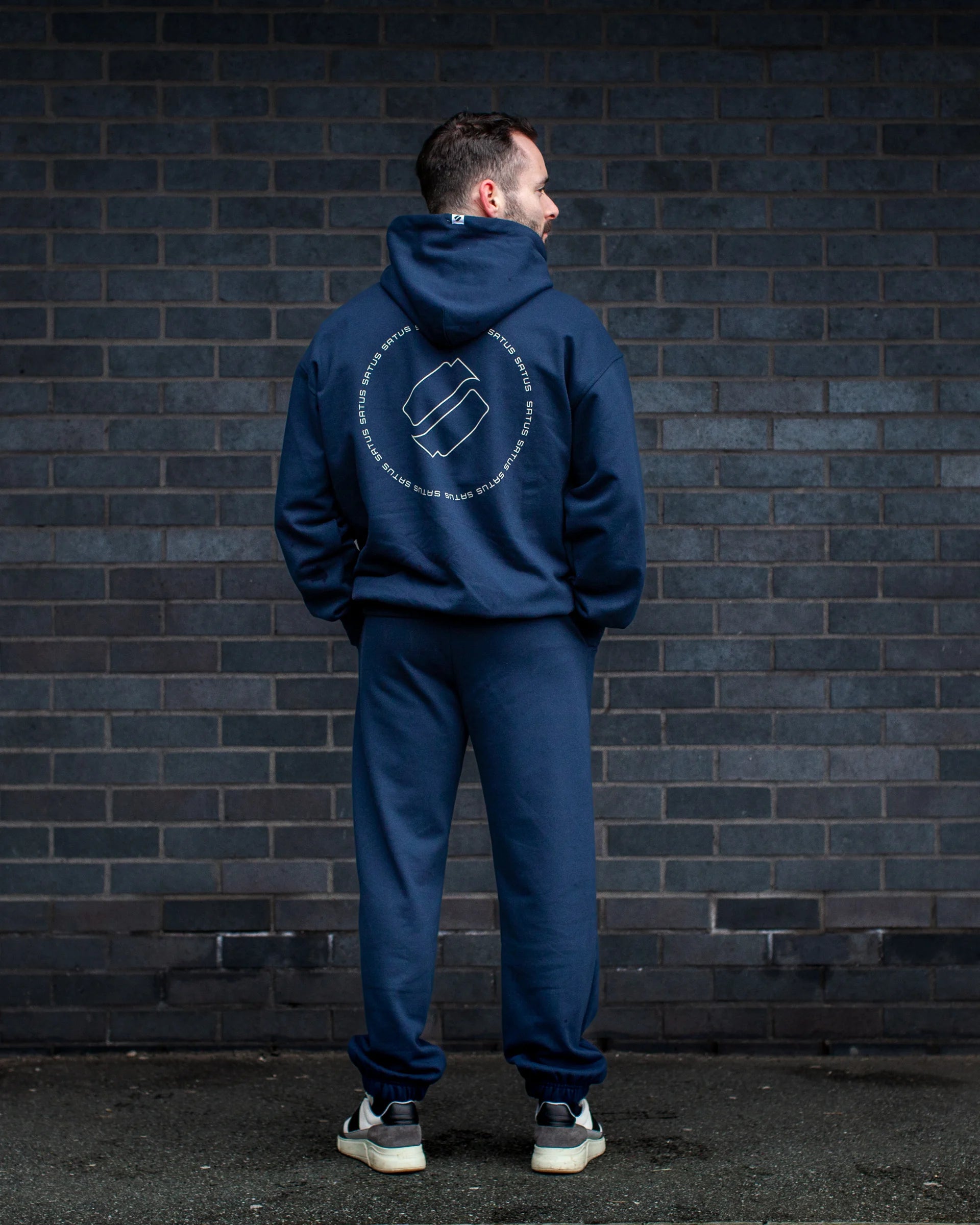 Person wearing a blue hoodie and joggers with a logo on the back, standing against a dark brick wall.