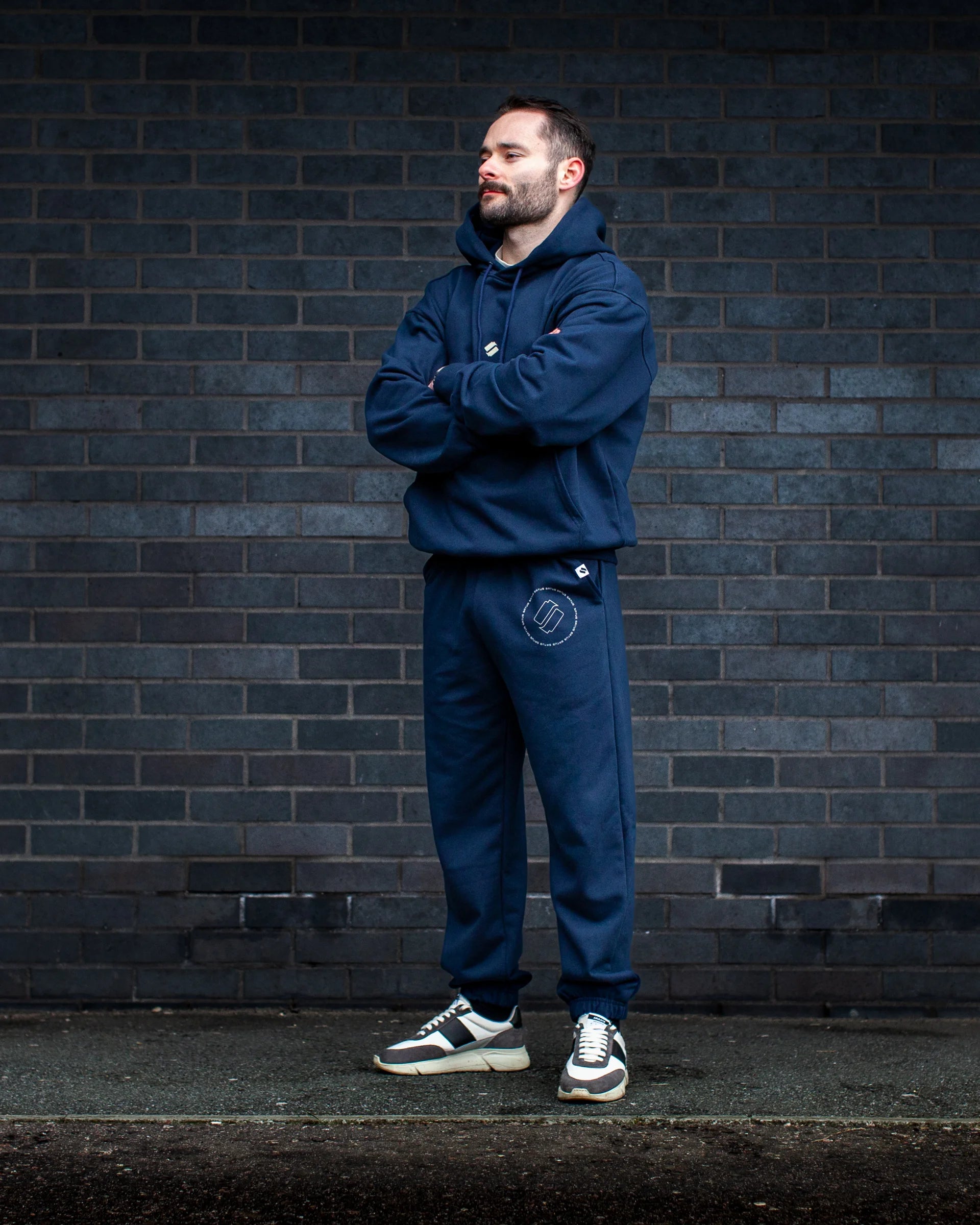 Man wearing a navy tracksuit standing against a dark brick wall.