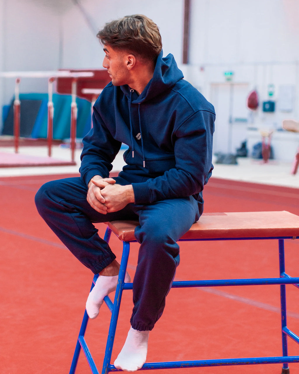 Person sitting on a platform in an indoor sports facility wearing blue hoodie and joggers