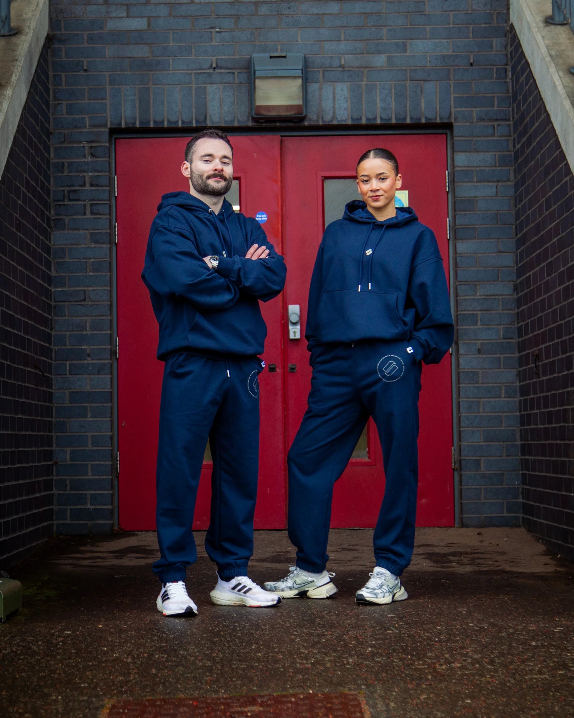Two people wearing matching blue tracksuits standing in front of a red door.