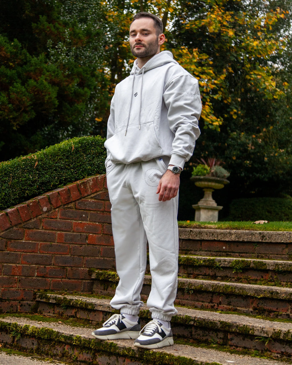Man wearing a light grey heavyweight hoodie hoodie and joggers standing on brick steps with greenery in the background
