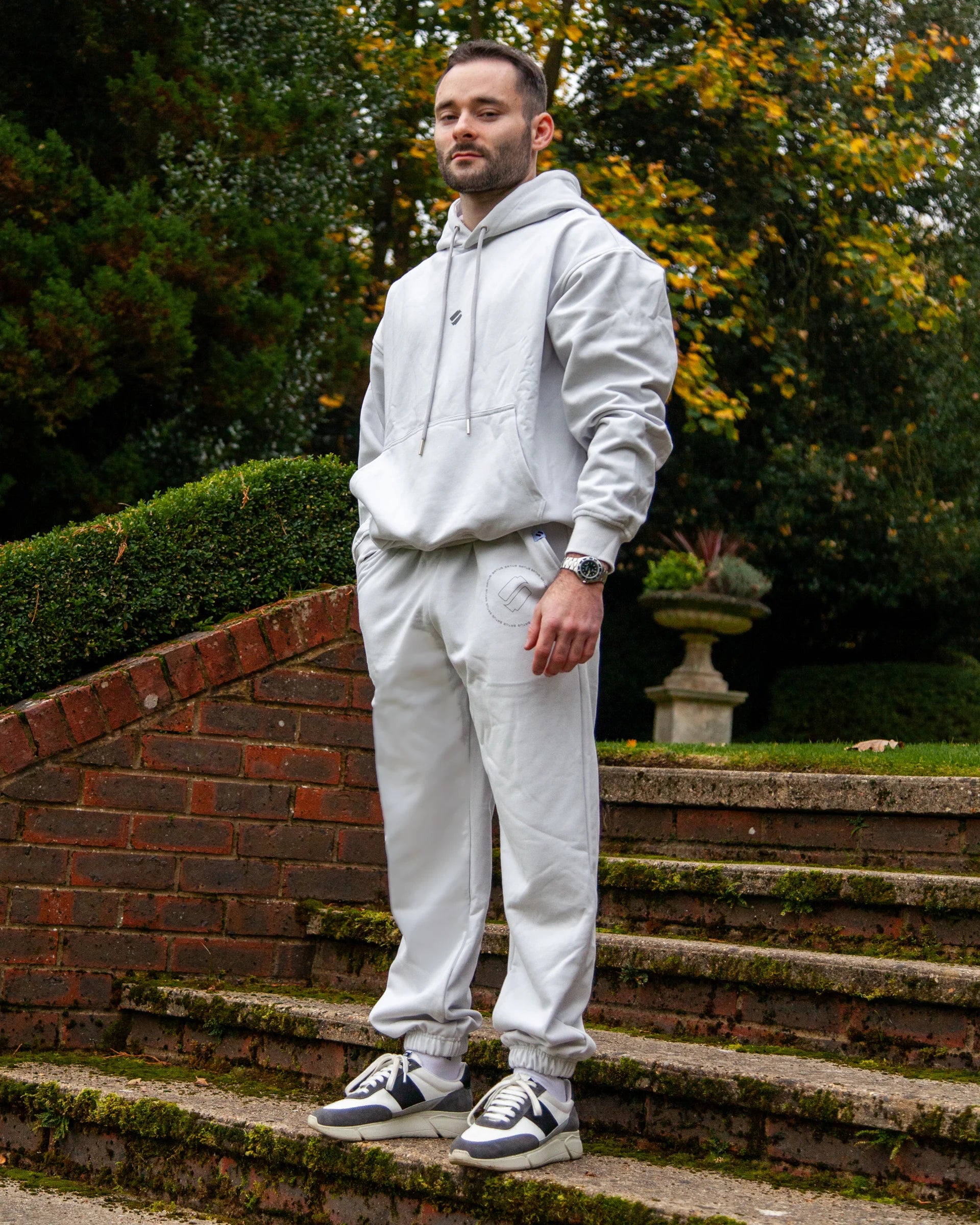Man wearing a light grey heavyweight hoodie hoodie and joggers standing on brick steps with greenery in the background