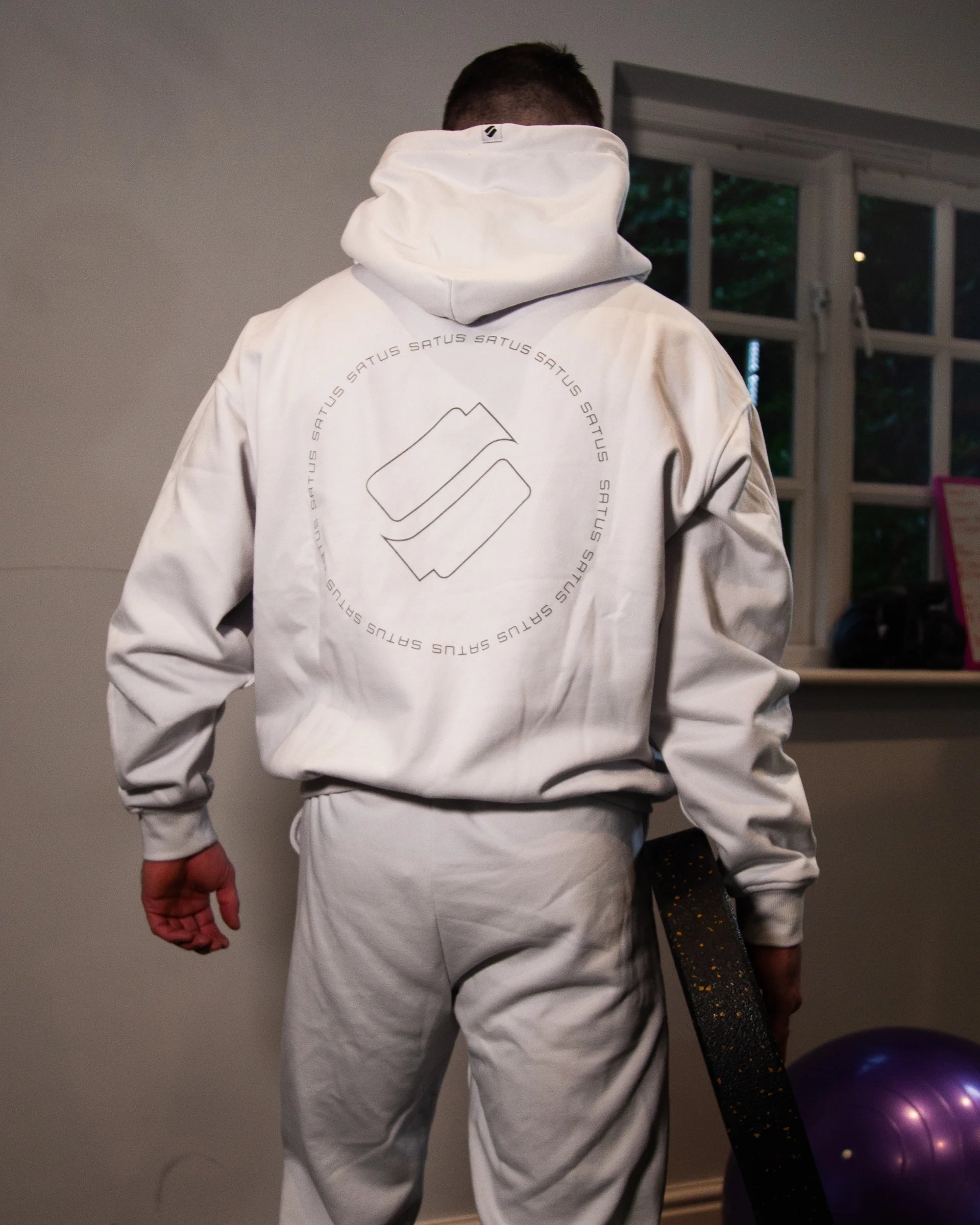 Person wearing a white hoodie with a logo on the back, standing in a home gym with a weight