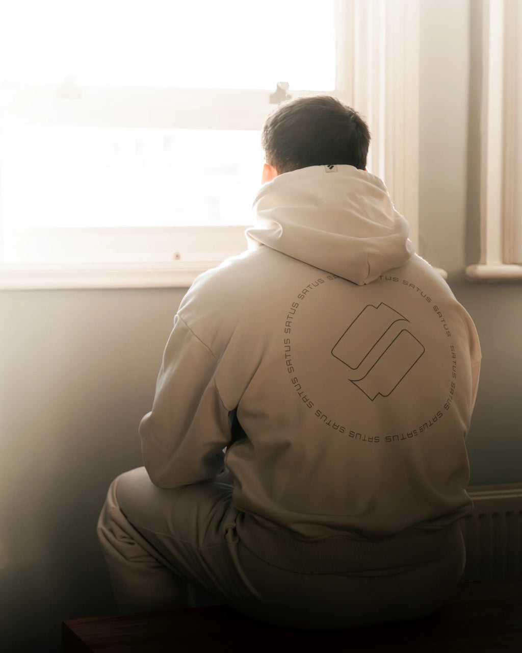 Person wearing a light grey hoodie with a logo sitting by a window looking out at the view