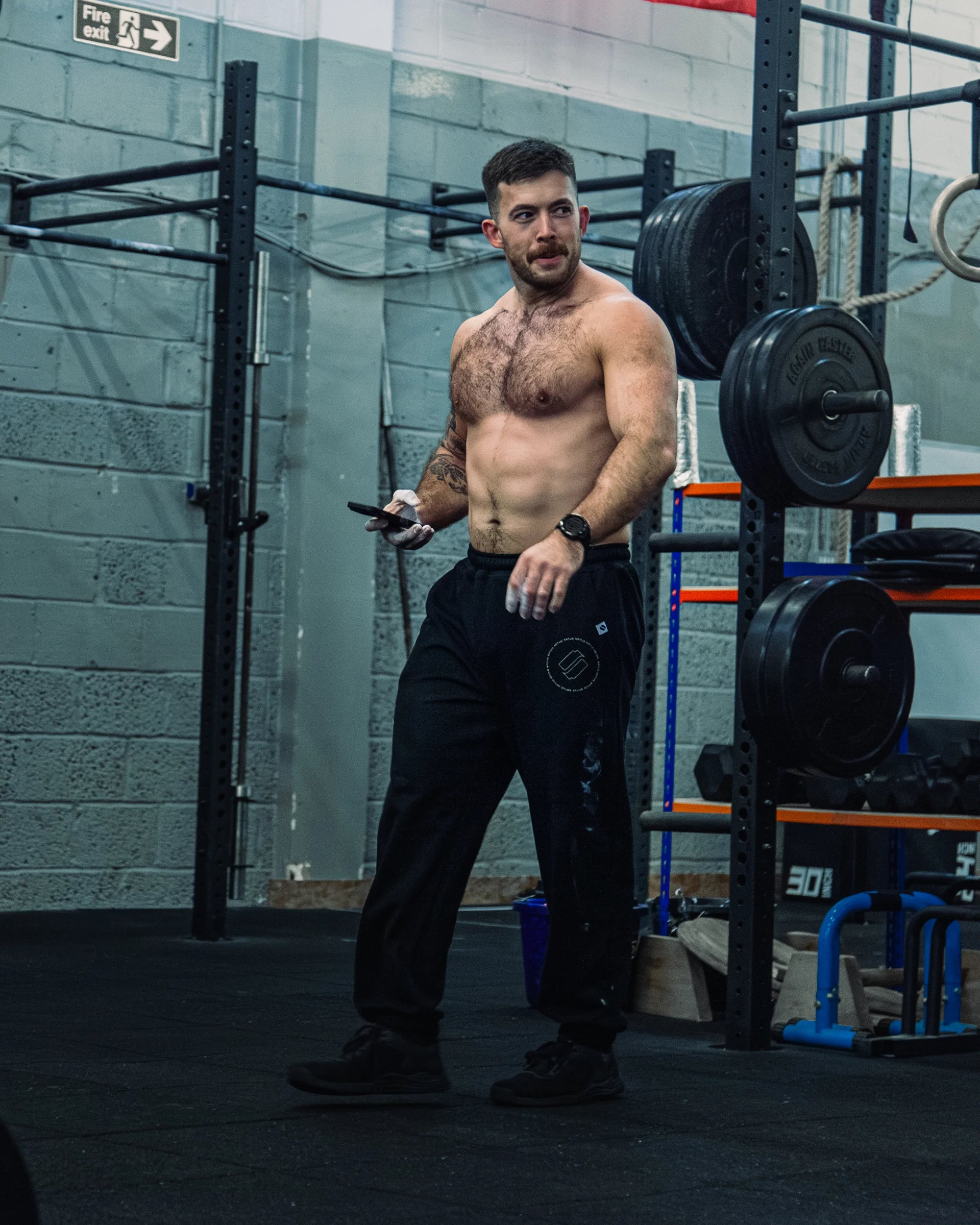 Man standing in a gym holding a smartphone with weightlifting equipment in the background wearing black joggers