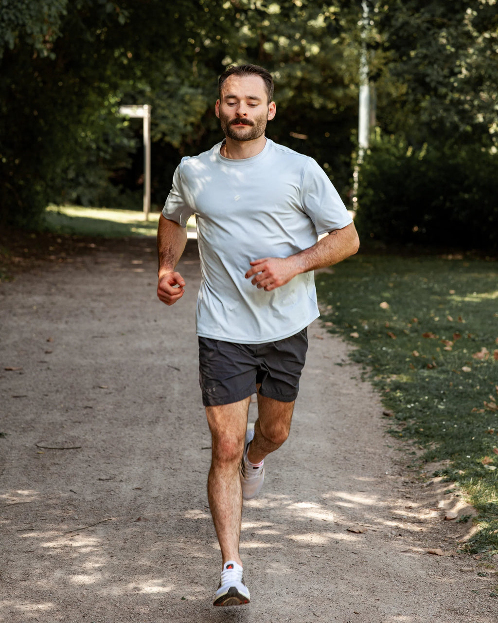 Man running on a path in a park with trees and greenery in the background wearing grey training shorts and blue training tee