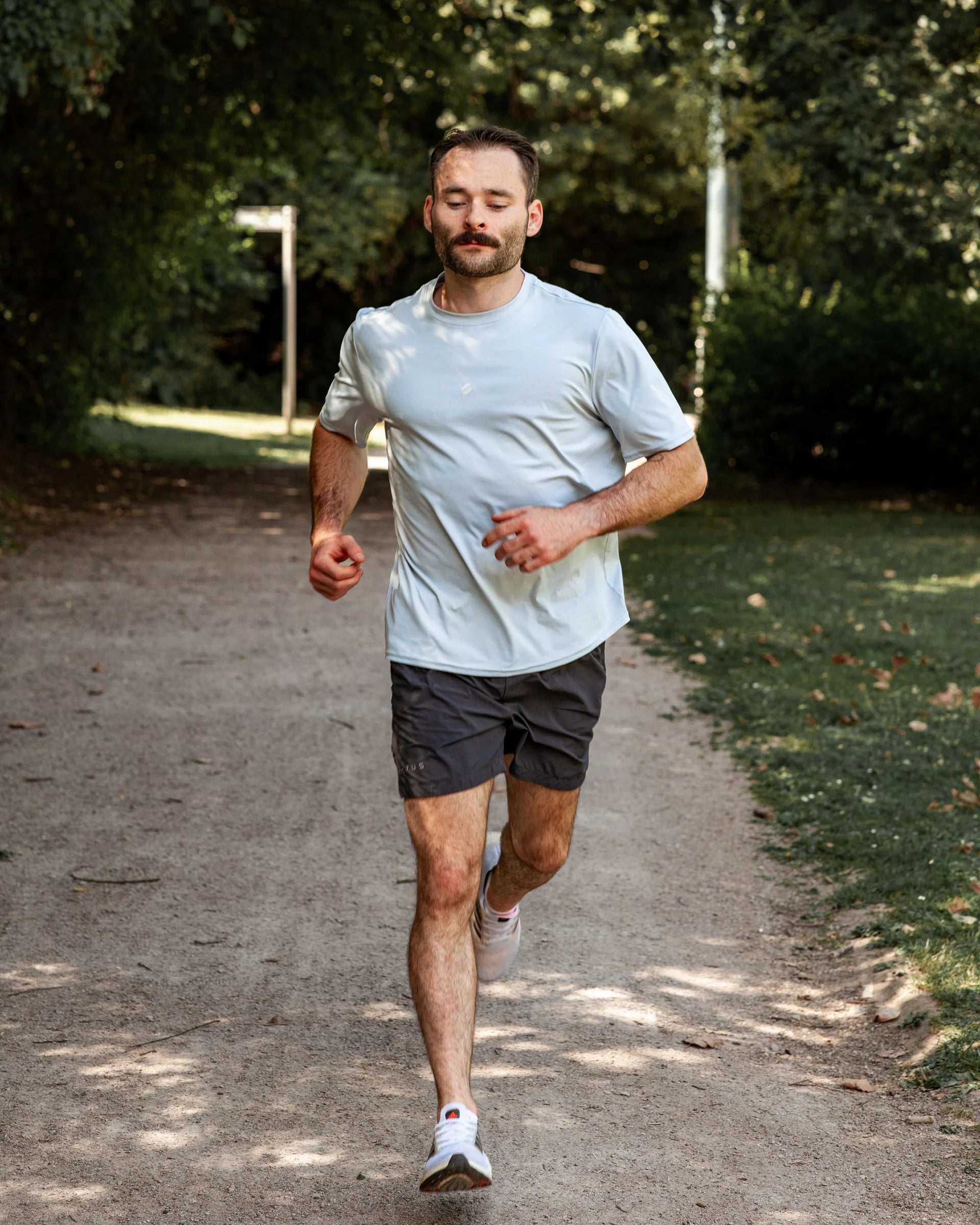 Man running on a path in a park with trees and greenery in the background wearing grey training shorts and blue training tee
