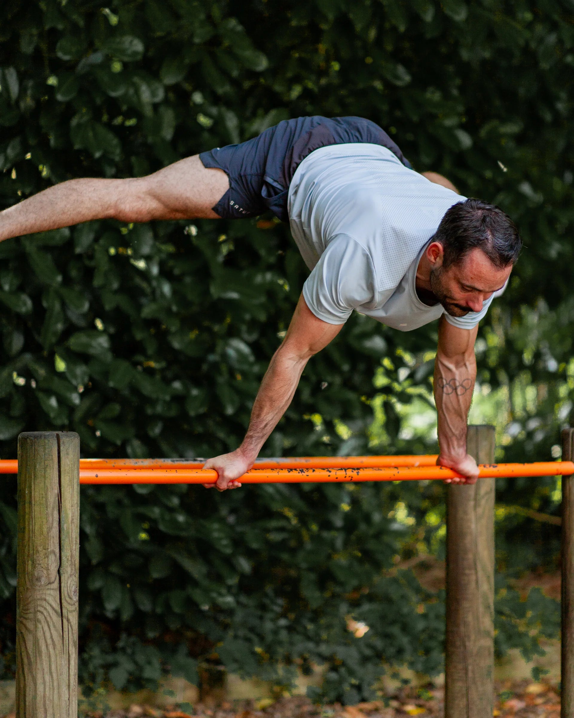 Man performing calisthenics on a horizontal bar outdoors with greenery in the background wearing grey training shorts and blue sports tee