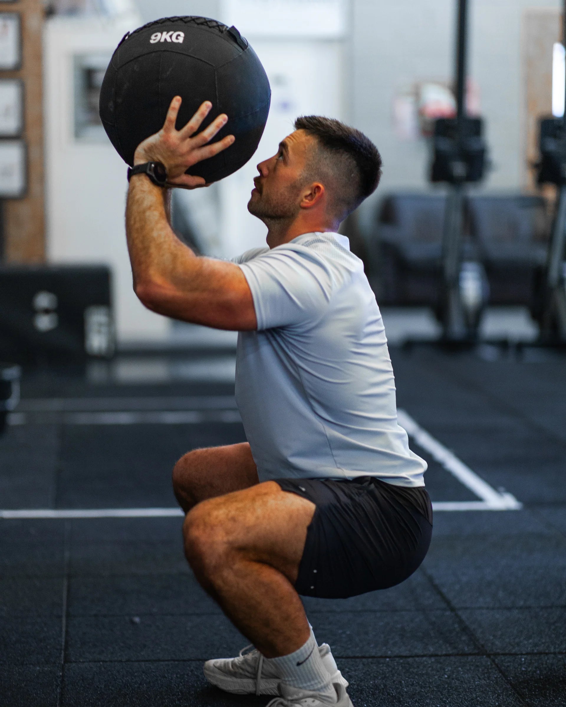 Man performing a squat exercise with a medicine ball in a gym setting wearing training shorts and a sports t-shirt