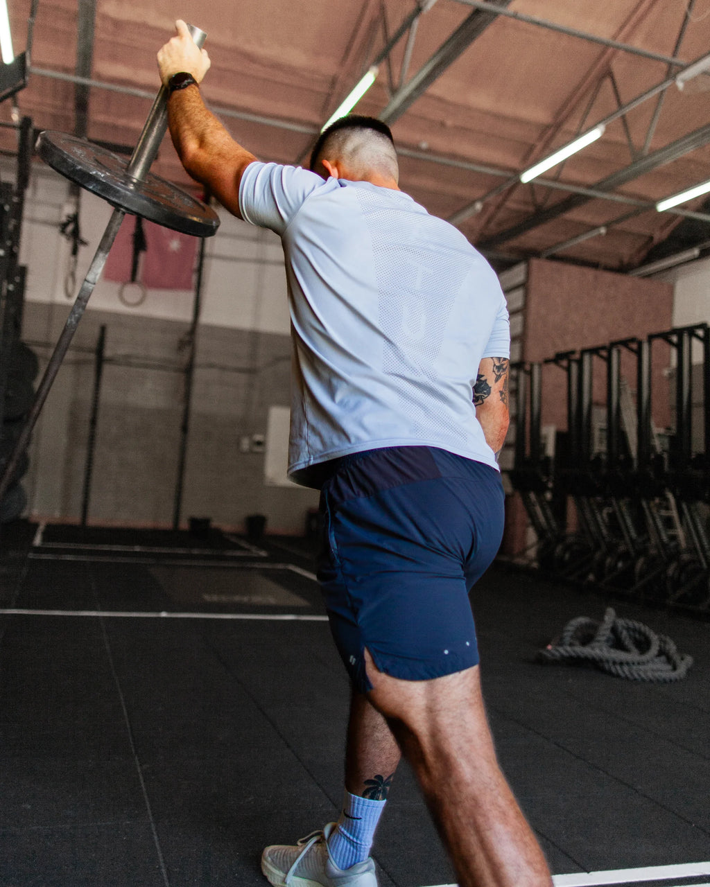 Man lifting a barbell in a gym setting wearing blue perforated tee and satus training shorts