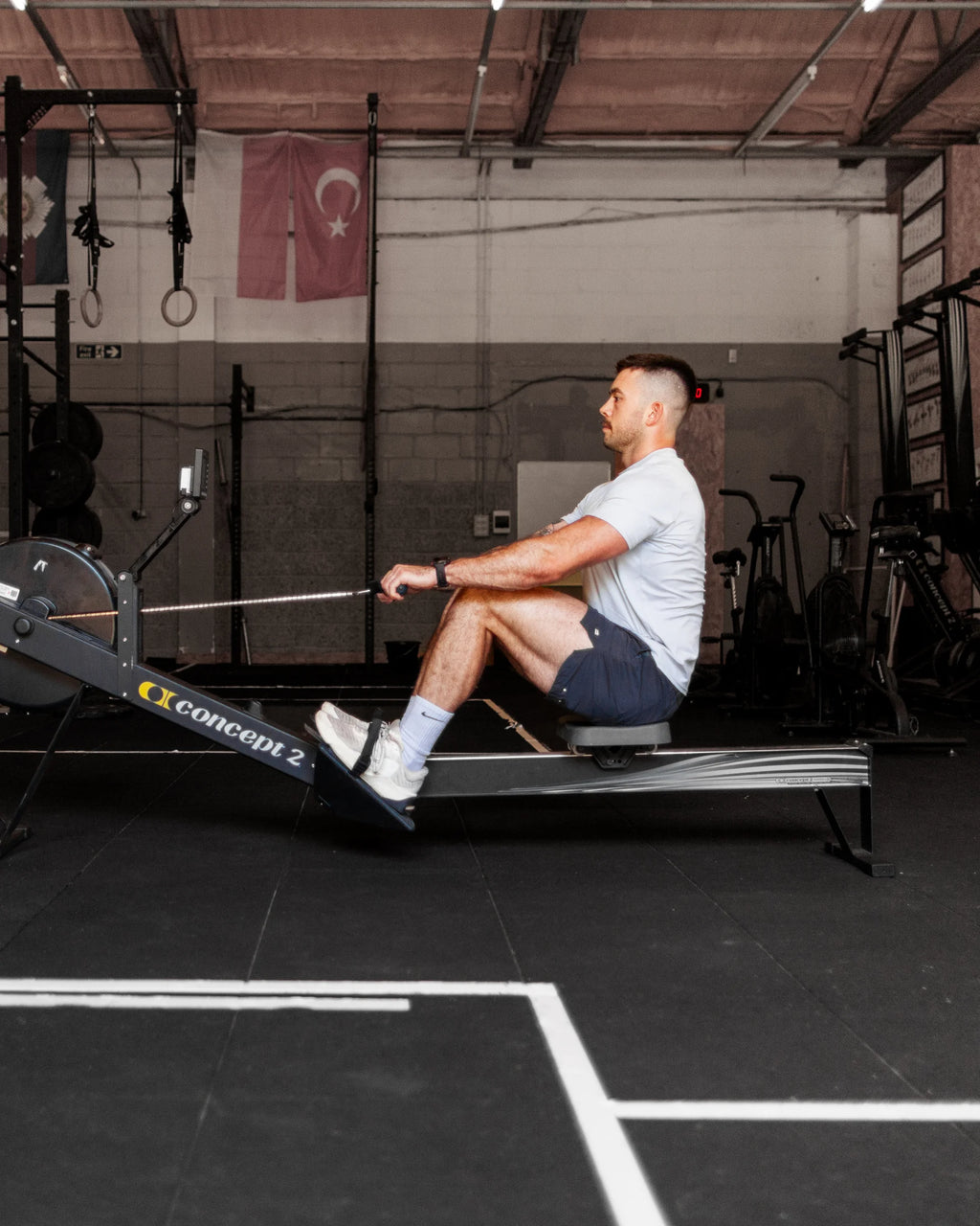Man using a rowing machine in a crossfit gym setting wearing training shorts and sports tee