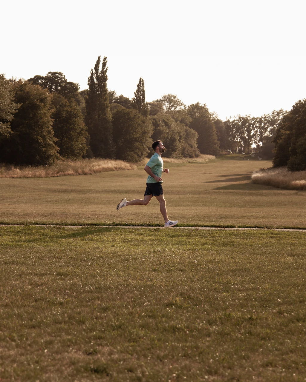 Msn running on a grassy field with trees in the background wearing green running top and training shorts